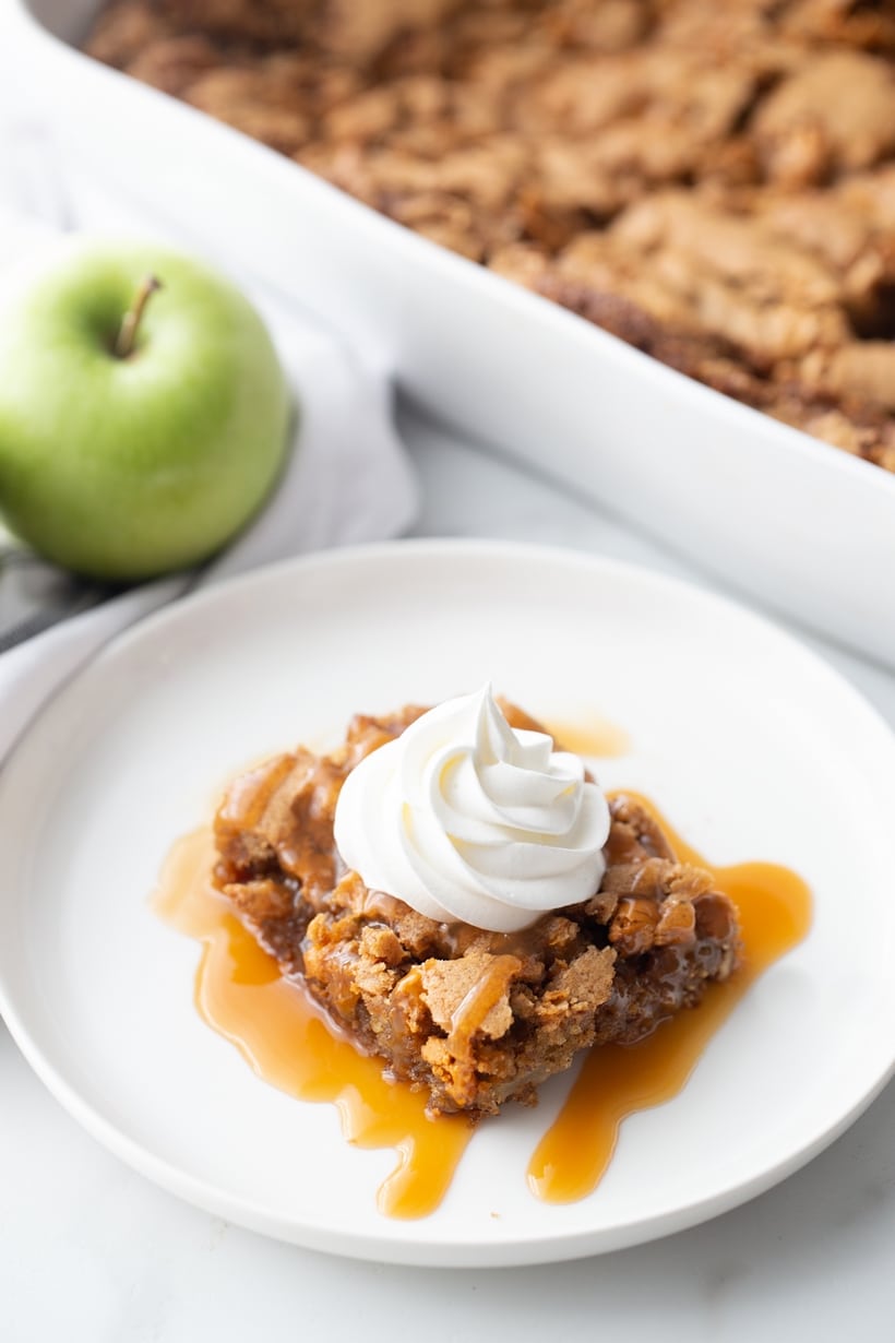 This light and airy apple cake filled with chunks of fresh apples and butterscotch morsels is the ultimate fall season dessert. Each slice finished with a drizzle of caramel and a dollop of whipped cream is decadence you need to experience. Apple cake on a plate with whipped cream.