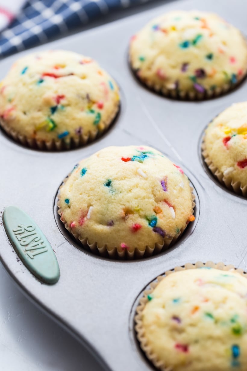 Unfrosted firecracker cupcakes showing rainbow specks inside. Freshly baked pop rock cupcakes cooling in a muffin tin before topping.