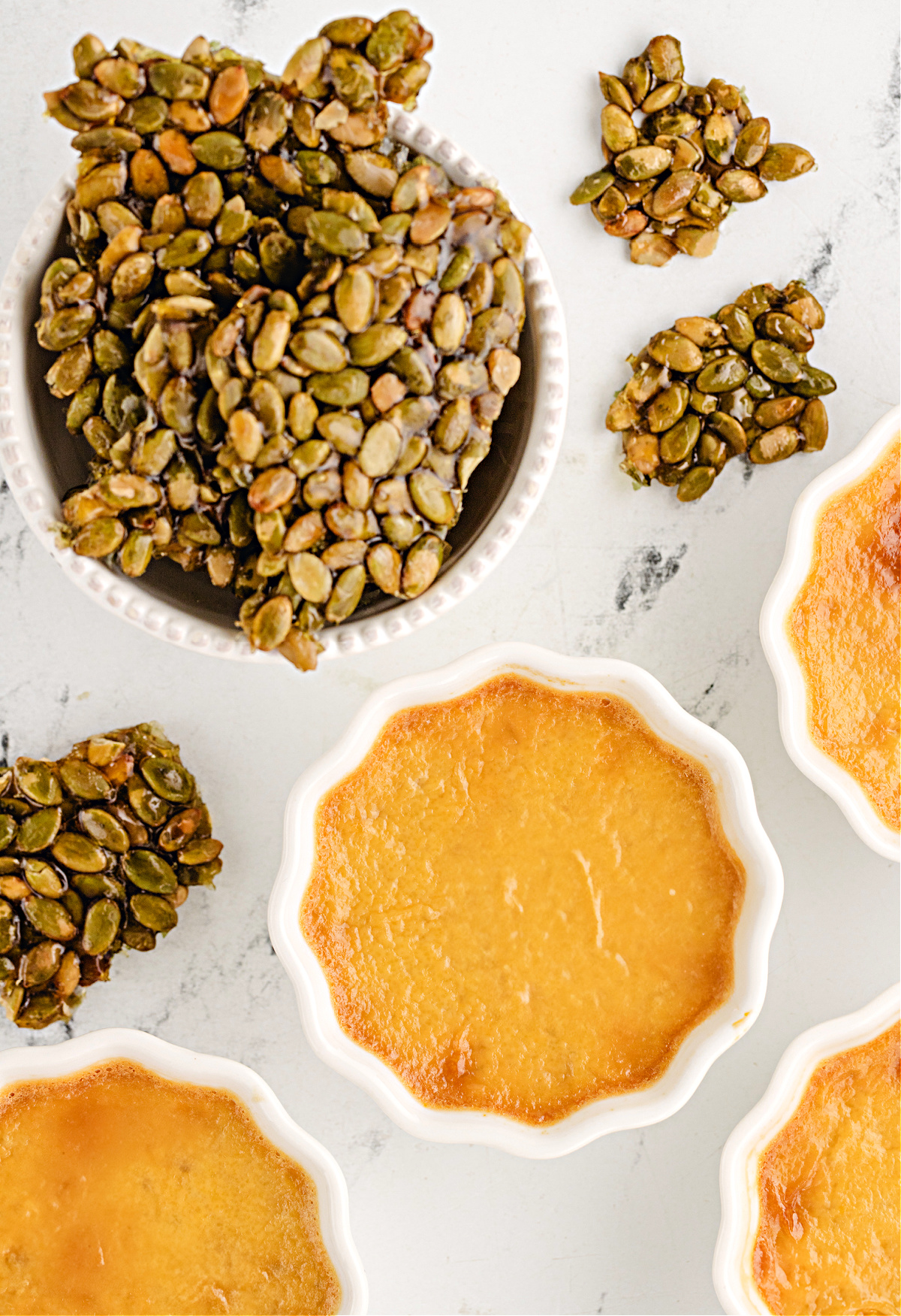 Pepita brittle cooling next to baked pumpkin chai custards, highlighting the crackly caramel contrast to the smooth pots de crème.