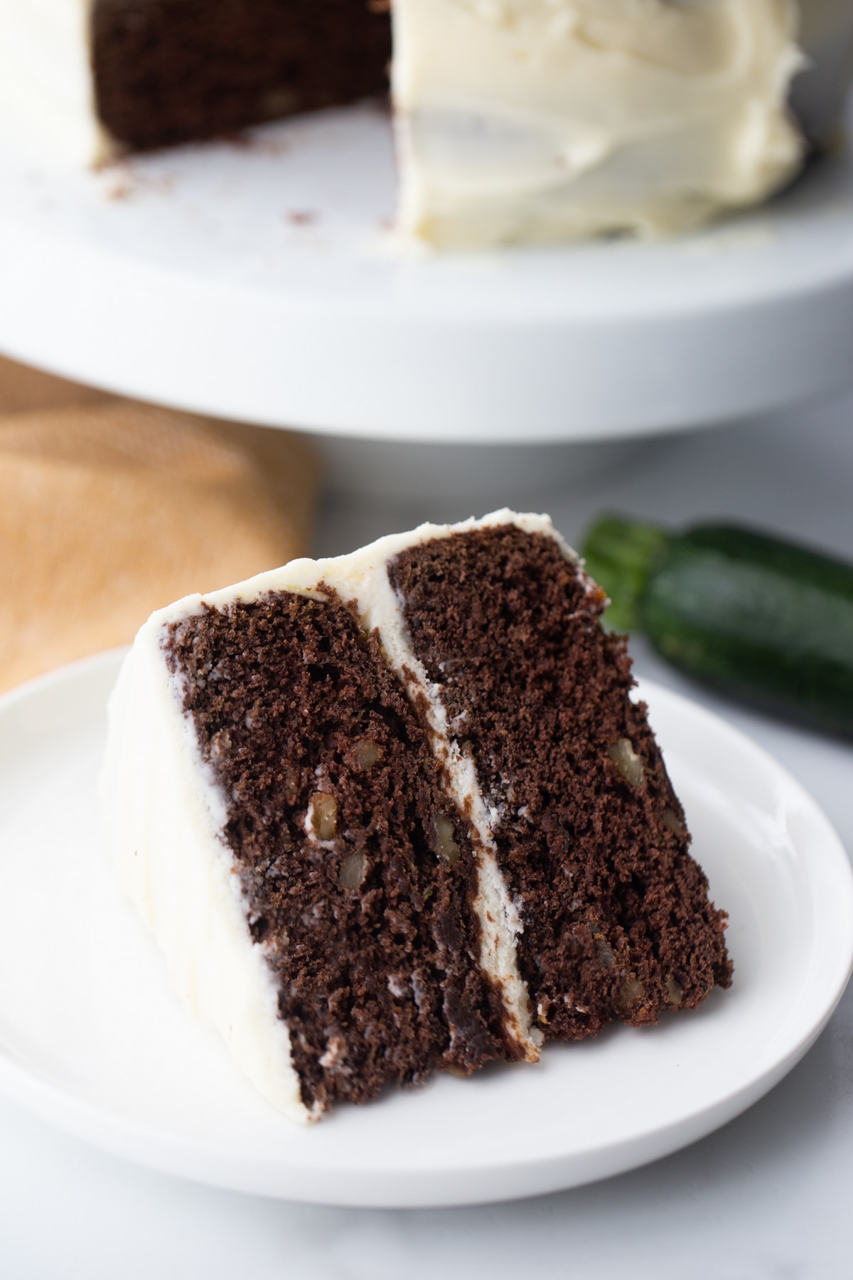 Slice of moist chocolate zucchini cake with cream cheese frosting, showing walnuts in the cake layers and zucchini in the background.