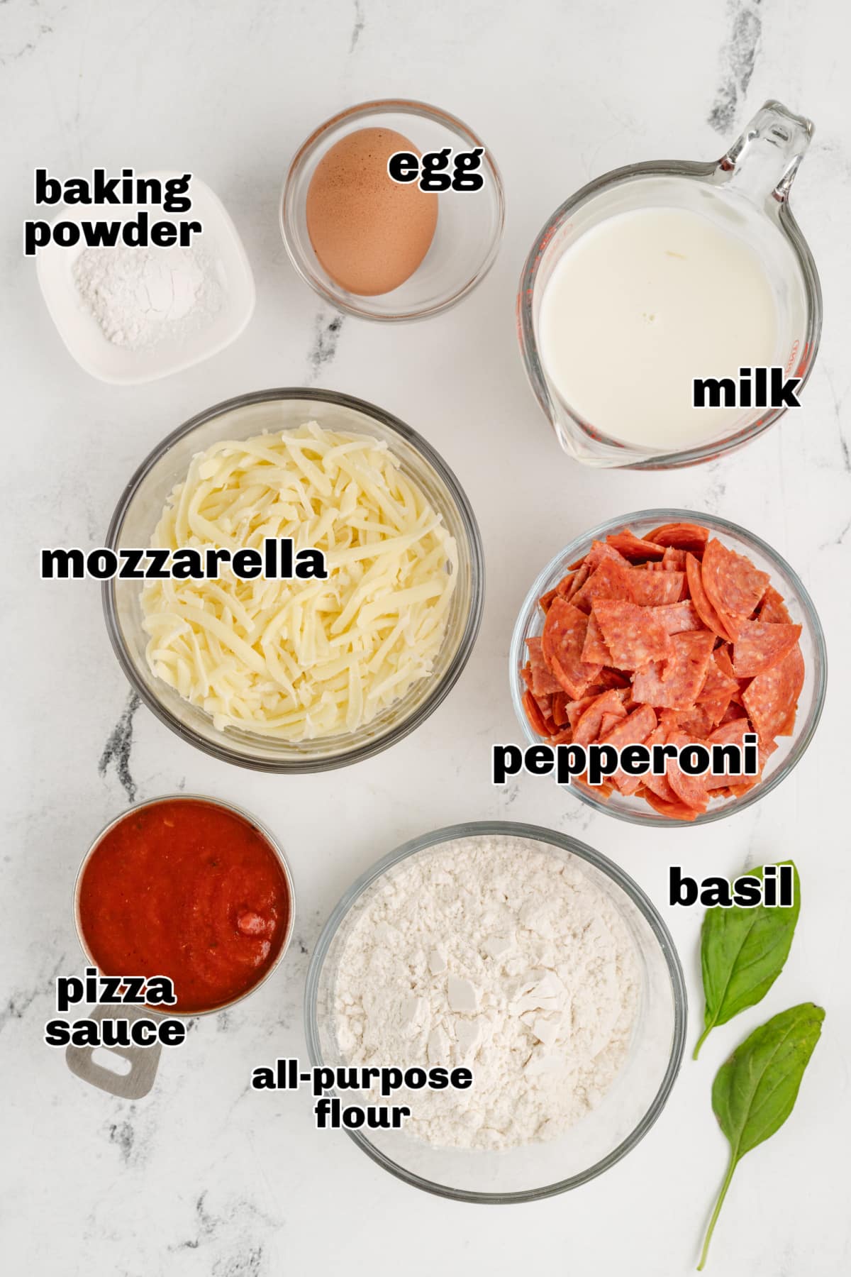 All the ingredients for the pepperoni pizza puffs displayed on a countertop, including flour, milk, egg, mozzarella, pepperoni, and seasonings.
