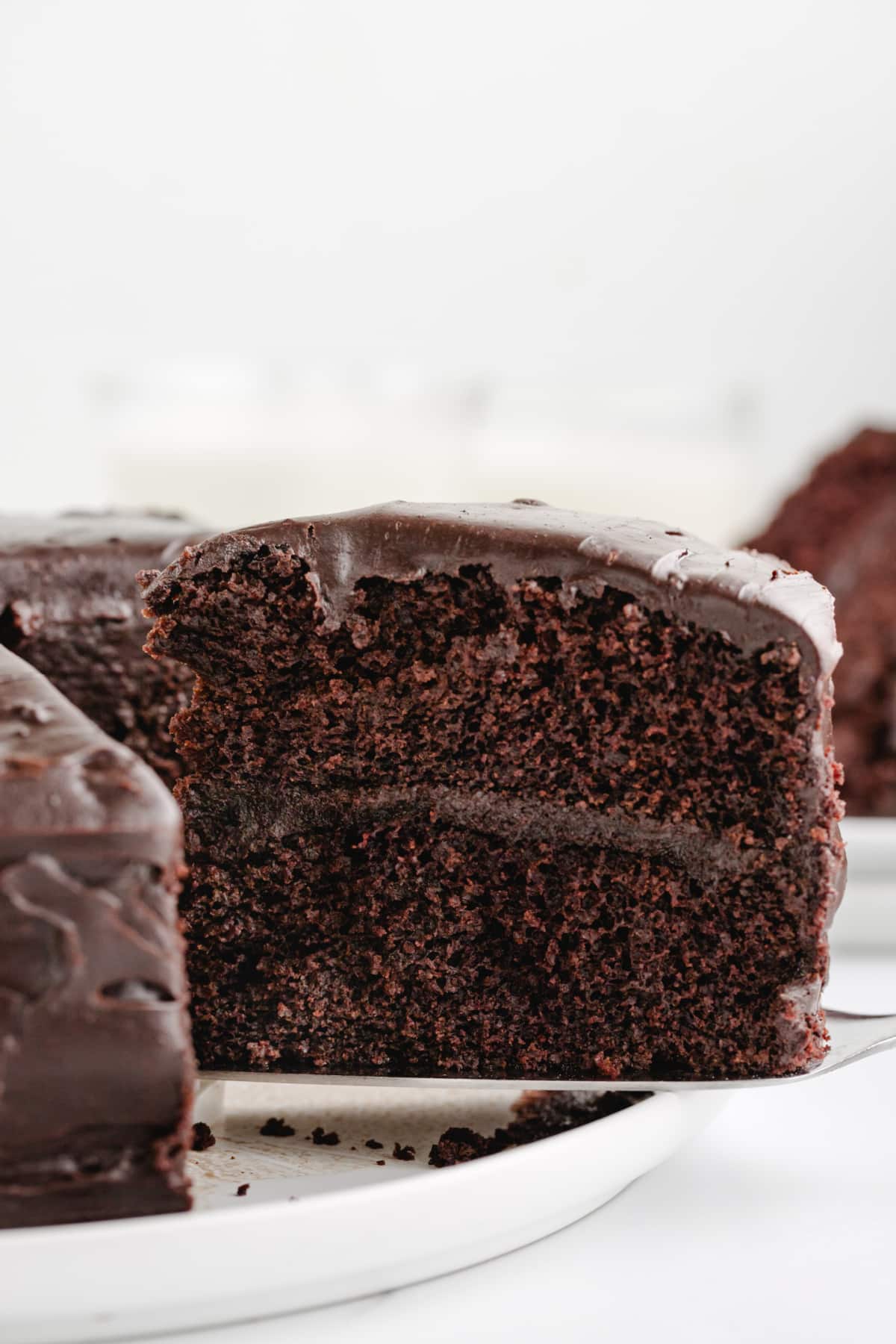 A layered slice of Old-Fashioned Chocolate Cake with Glossy Chocolate Icing Side angle of a sliced piece of chocolate cake, with layers of glossy chocolate icing between soft cake layers.