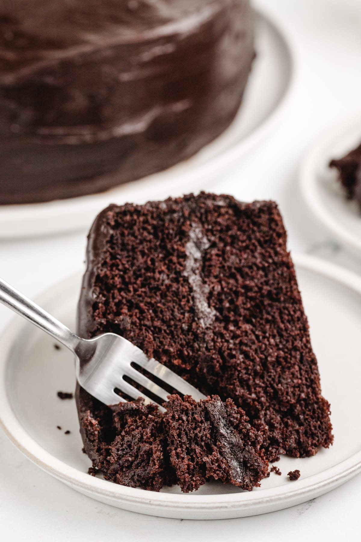 Old-Fashioned Chocolate Cake with Glossy Chocolate Icing being eaten with a fork A fork cutting into a slice of chocolate layer cake, highlighting the soft texture and thick chocolate icing.
