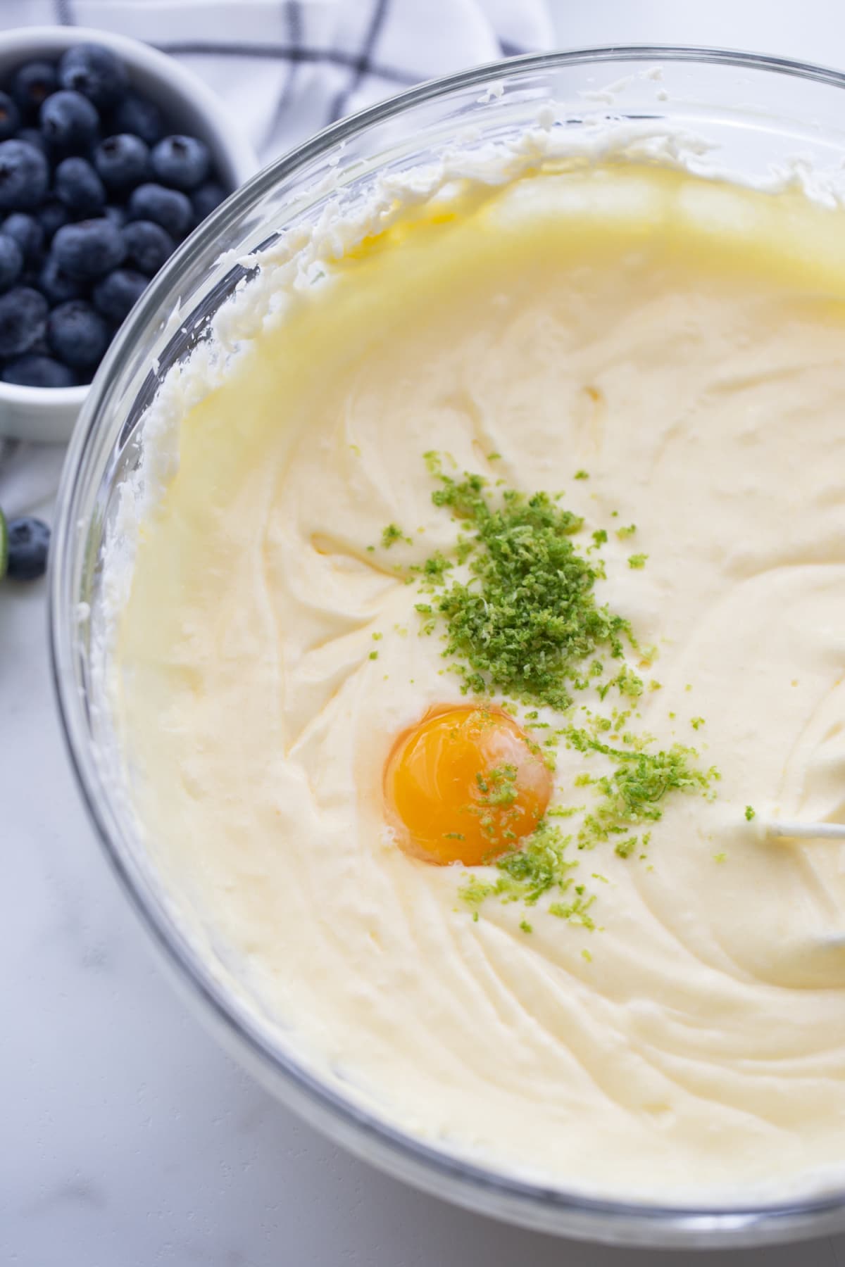 Blueberry-Lime Pound Cake batter Mixing bowl filled with pale batter, with a full egg yolk and fresh lime zest sitting on top before being mixed in.