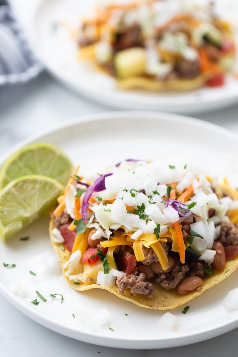 Two finished chipotle beef tostadas with cheese, slaw, and lime wedges