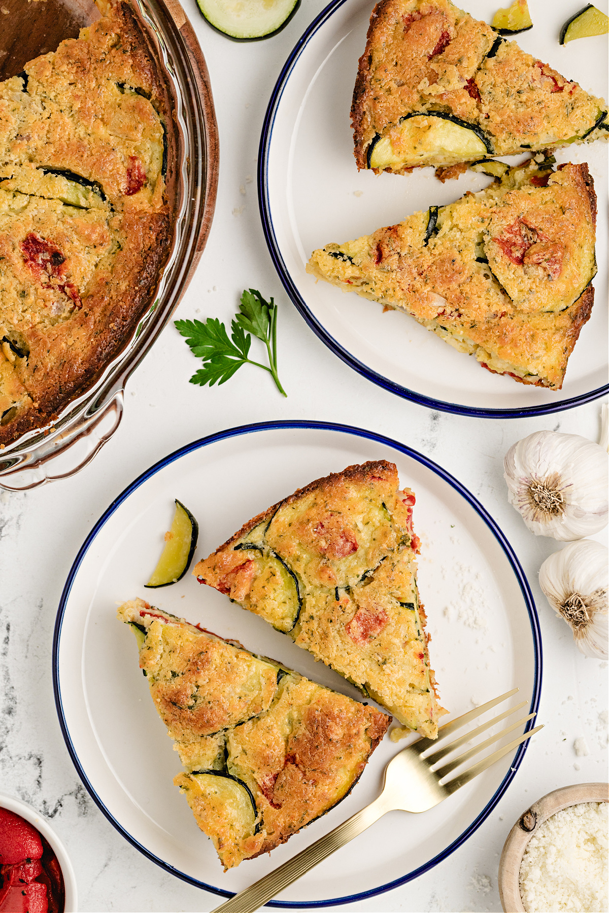 Easy zucchini pie with biscuit mix served for lunch Slices of savory zucchini pie on white plates with garlic, parsley, and roasted pepper on the side