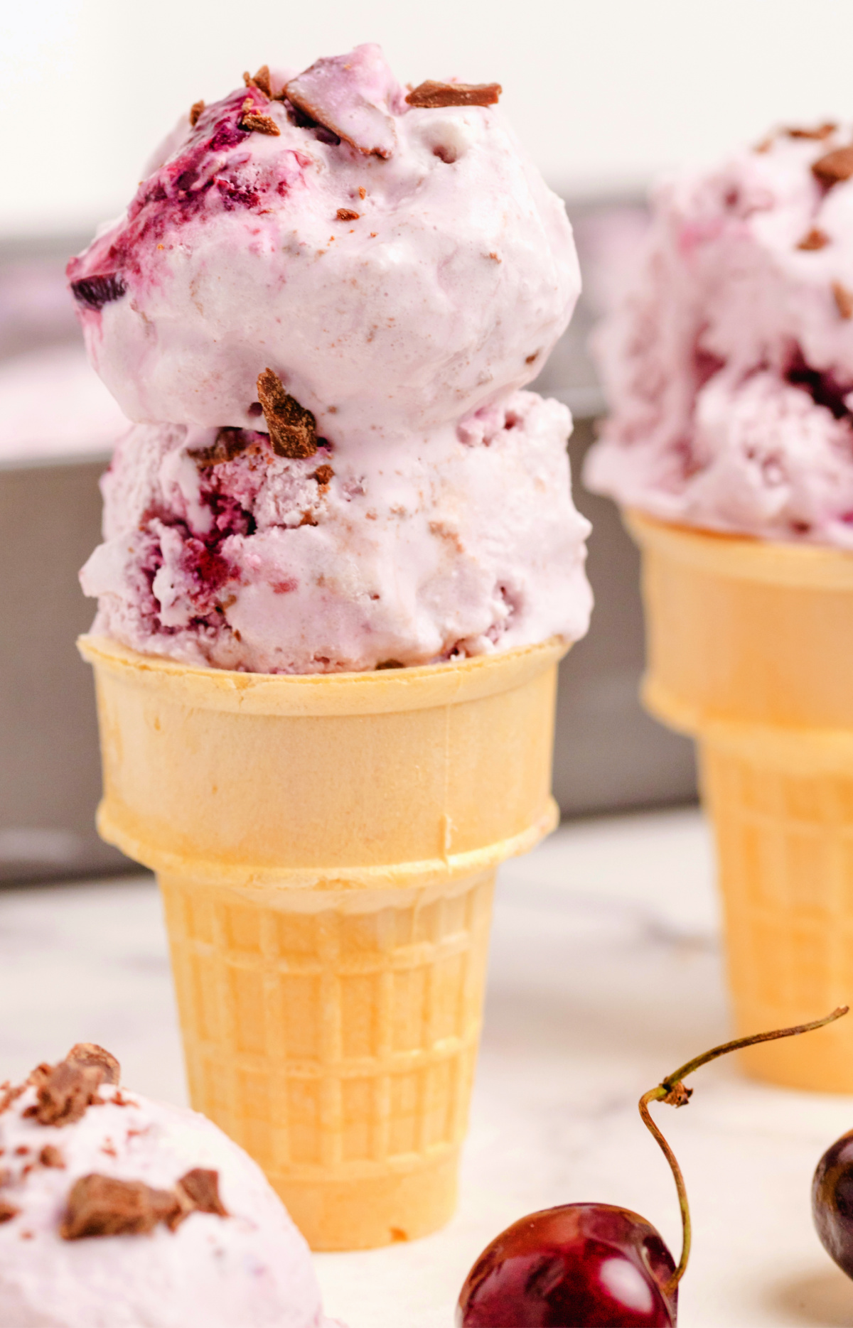 Cherry coconut chocolate chip ice cream served in a cone with chopped chocolate pieces, showing churned texture and visible fruit swirls.