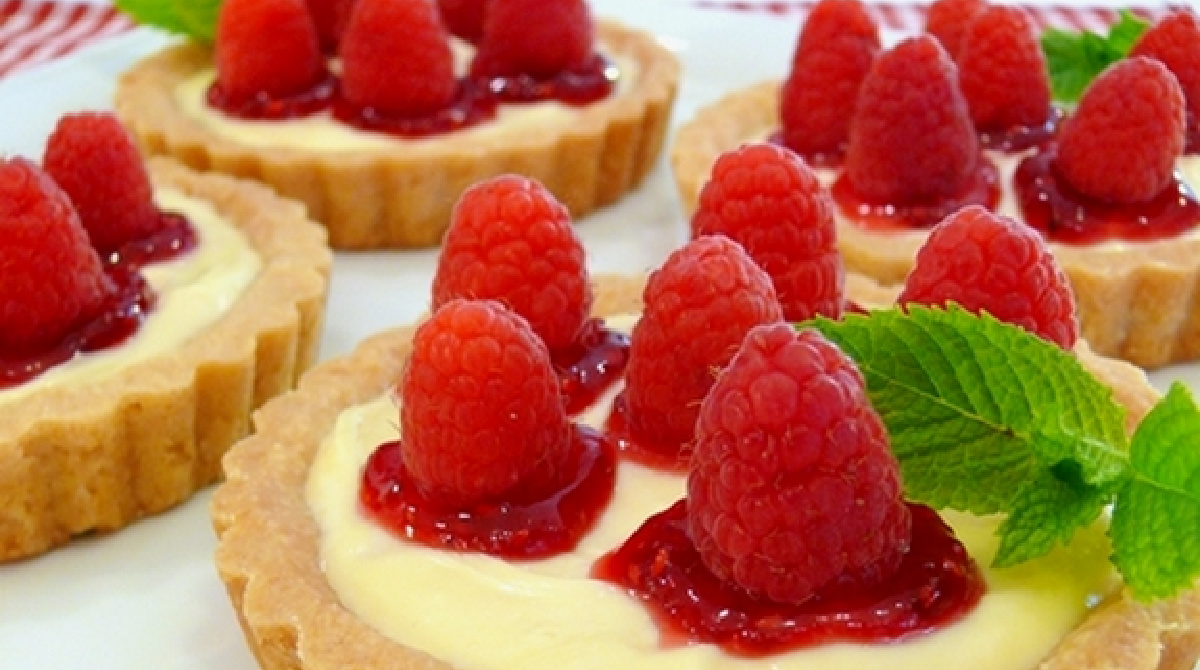 Homemade raspberry lemon tartlets topped with fresh raspberries and raspberry jam on a white serving platter.