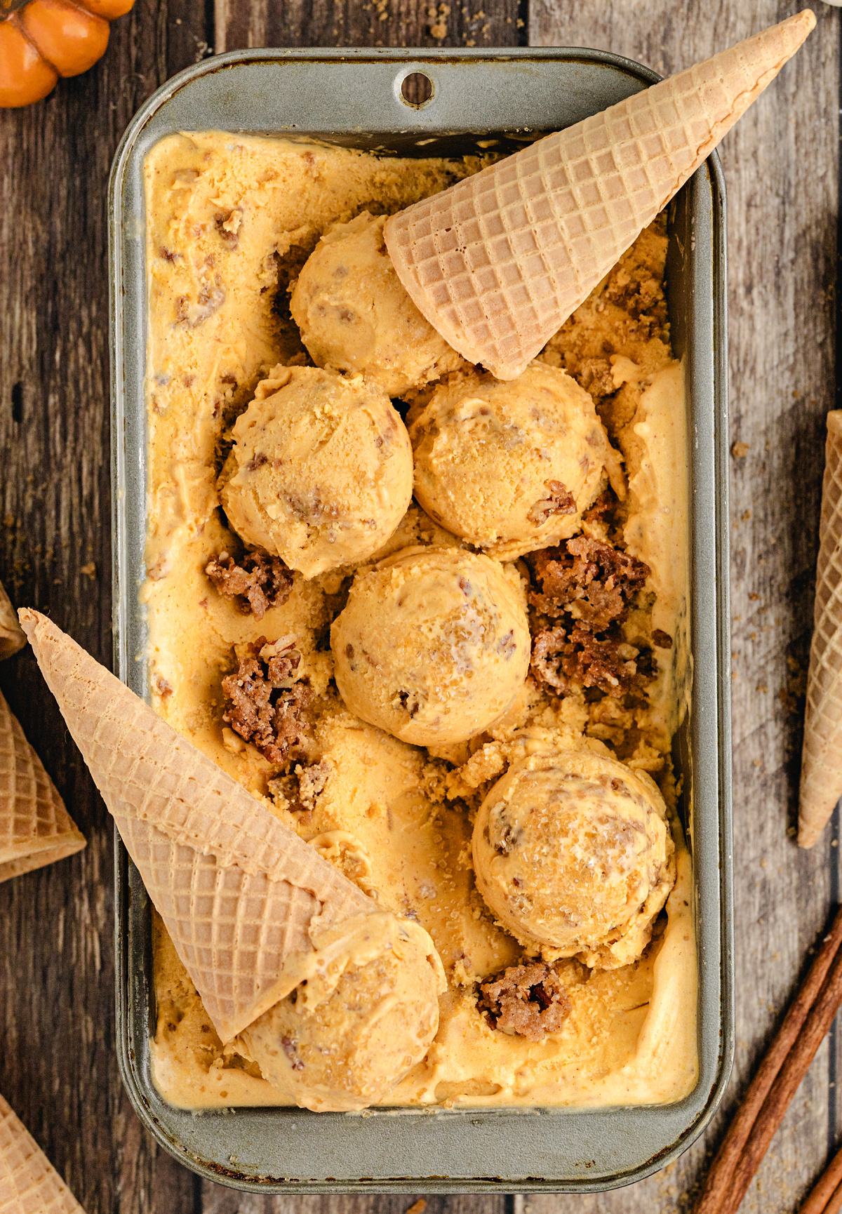 Scoops of Pumpkin Brown Sugar Crunch Ice Cream in a metal pan with waffle cones and baked streusel pieces, showing creamy texture and rich brown sugar color.
