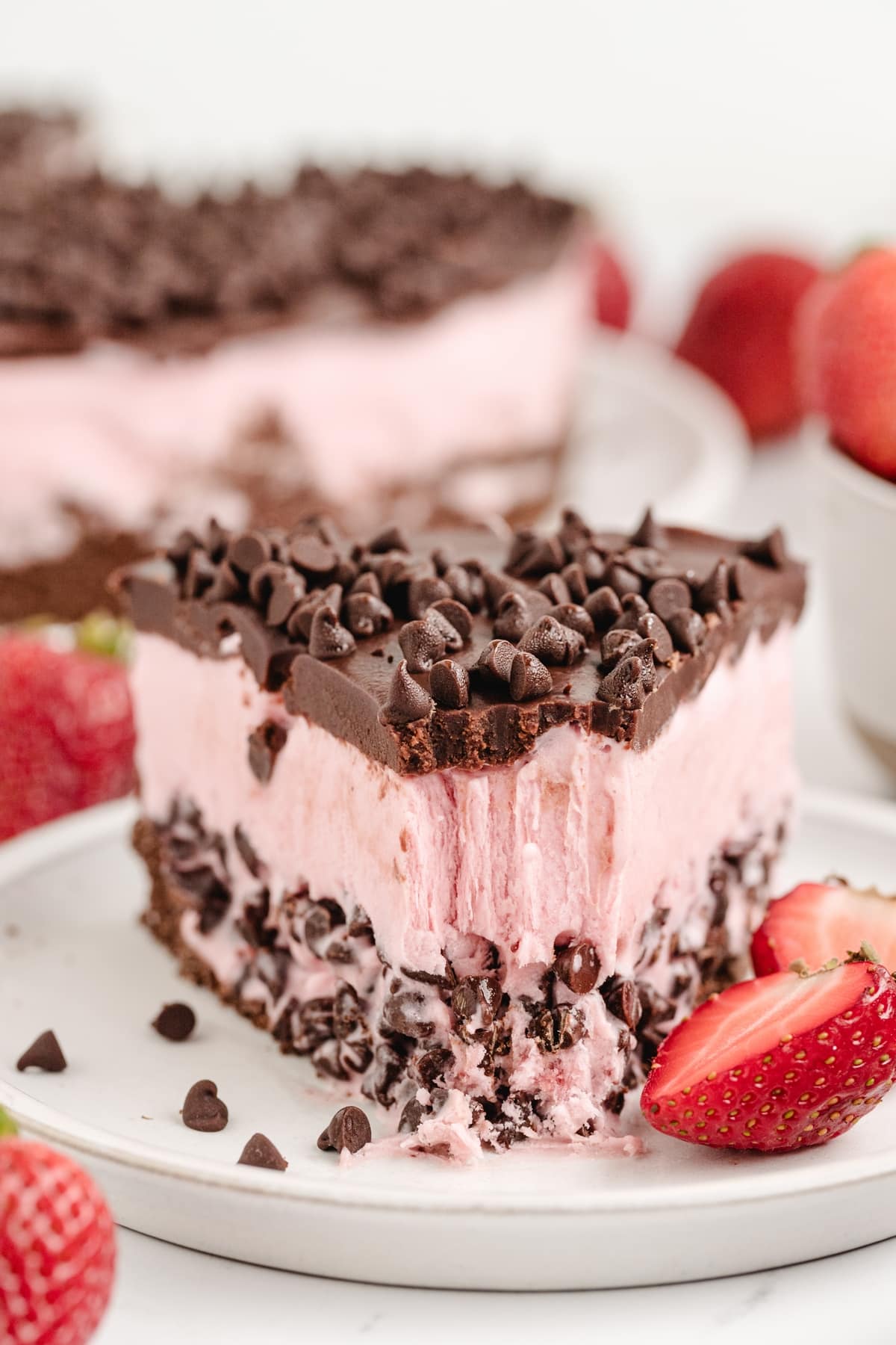 A partially eaten slice of strawberry chocolate chip ice cream cake on a plate, showing layers of chocolate graham cracker crust, creamy strawberry ice cream with mini chocolate chips, and a glossy chocolate ganache topping for Noble Pig