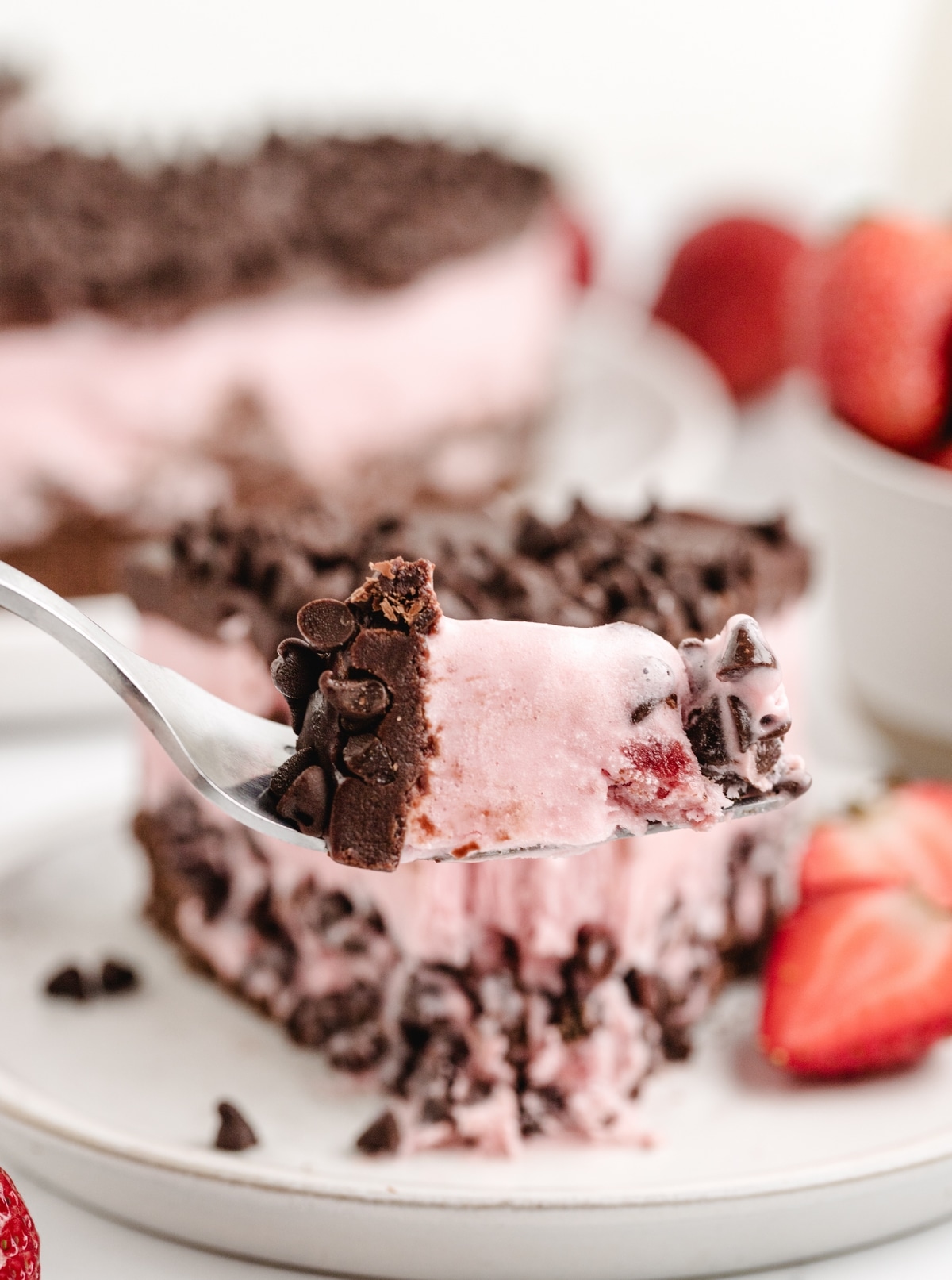 A fork lifting a bite of strawberry chocolate chip ice cream cake, showing the creamy ice cream and chocolate ganache with flecks of mini chocolate chips.