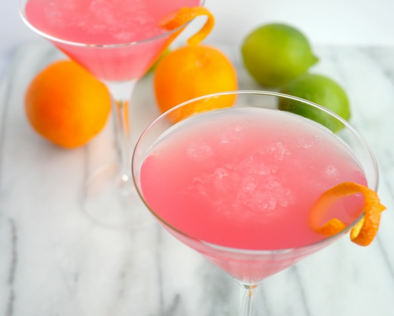 World's Best Cosmopolitan Cocktail in two martini glasses with limes and oranges in the background.