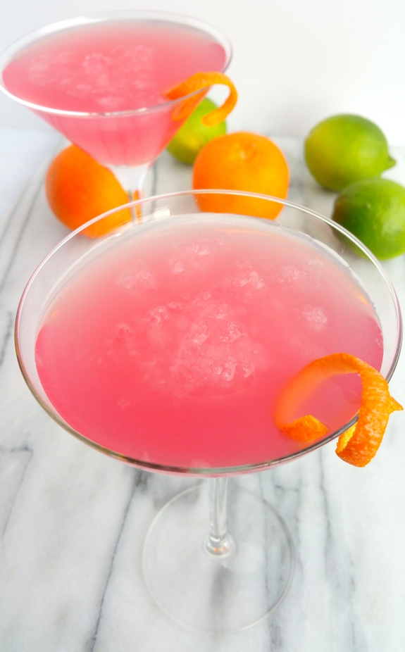 Two of the World's Best Cosmopolitan Cocktails in martini glasses with limes and oranges in the background.