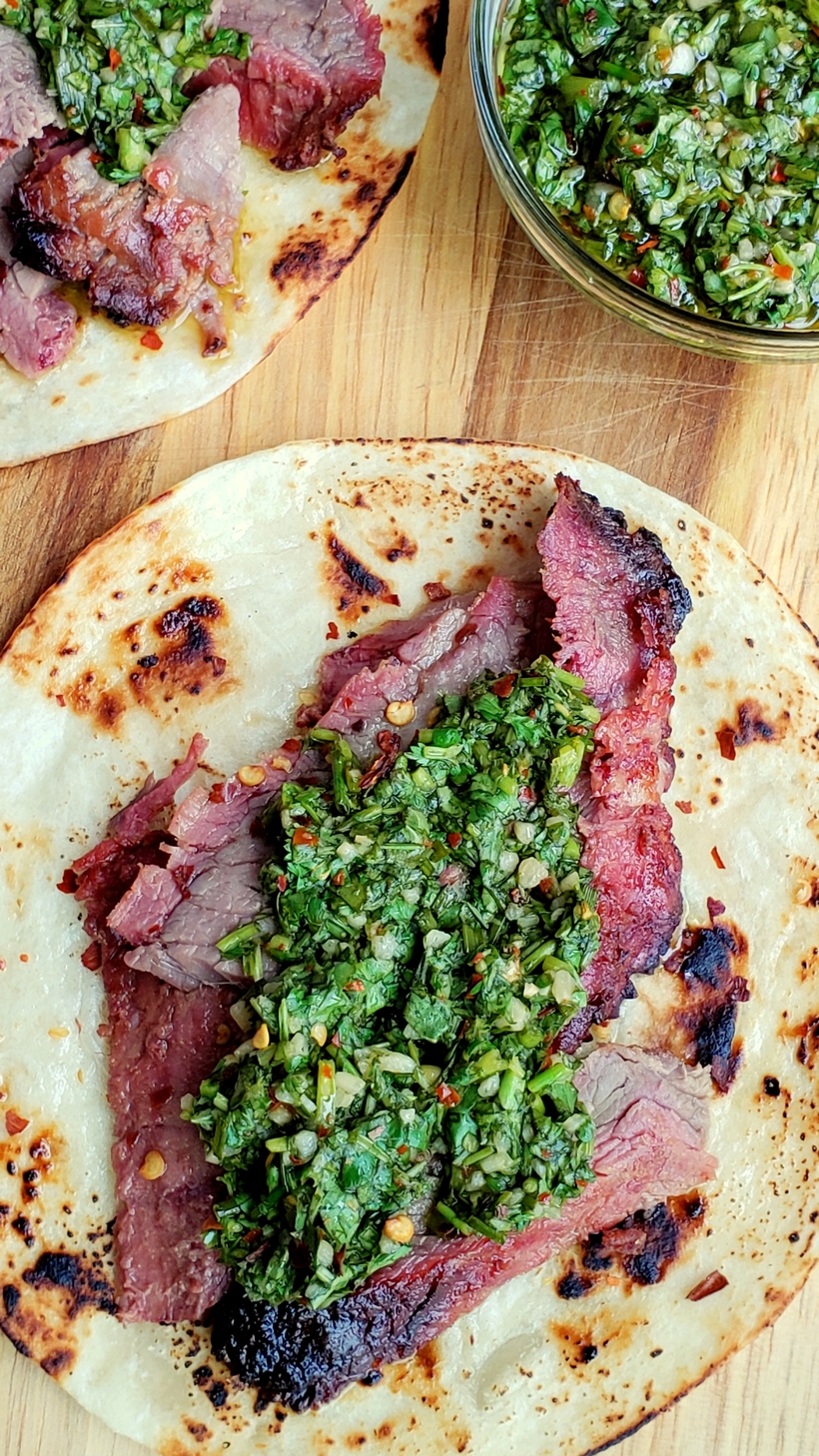 Make the best street food at home with these Flank Steak Tacos with Garlicky Cilantro Chimichurri. These tacos are essentially going to be your go-to summer, grilling meal. #noblepig #grilling #beef #flanksteak #chimichurri Overhead of Flank Steak Tacos with Garlicky Cilantro Chimichurri and a bowl of chimichurri.
