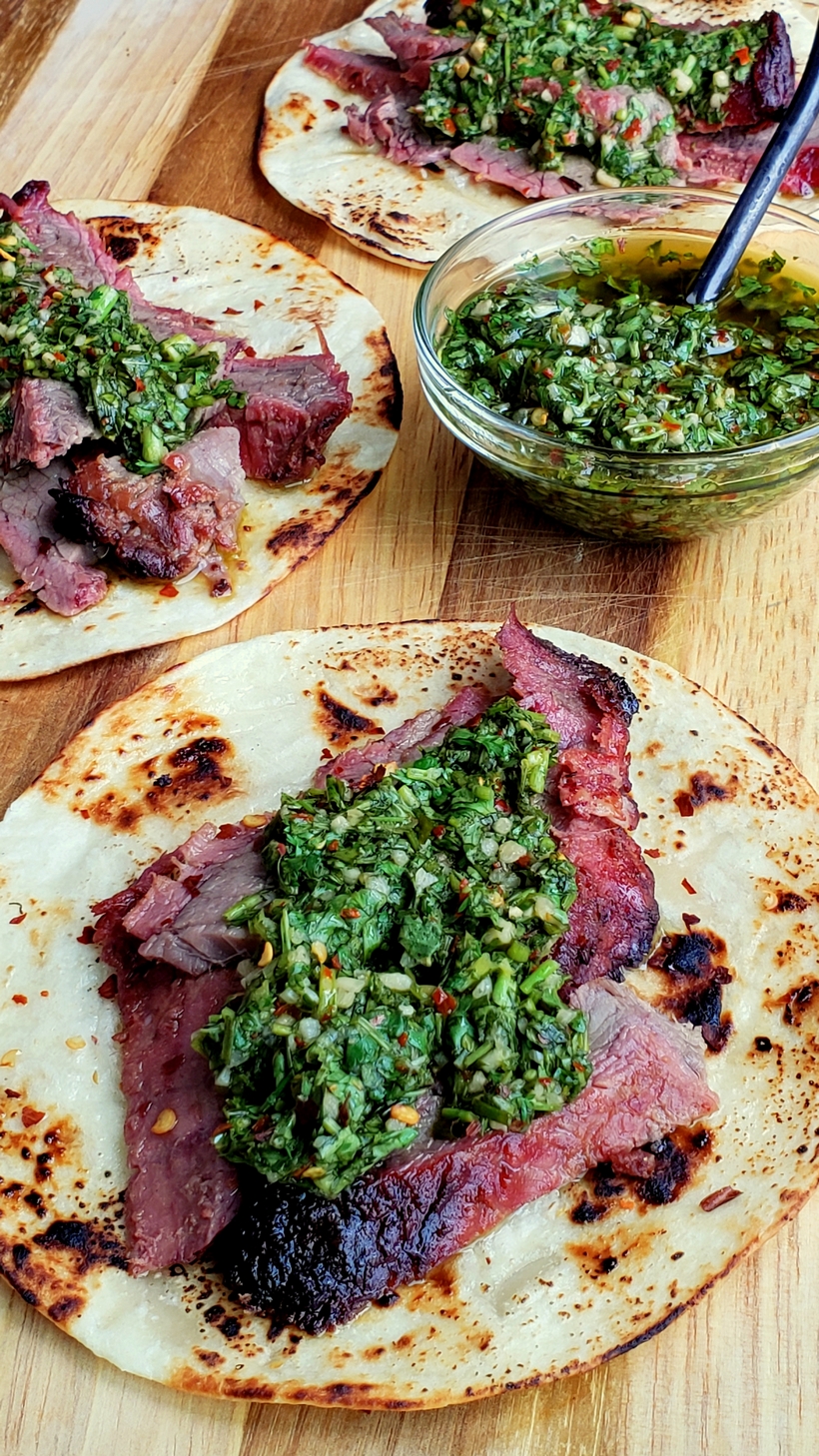 Make the best street food at home with these Flank Steak Tacos with Garlicky Cilantro Chimichurri. These tacos are essentially going to be your go-to summer, grilling meal. #noblepig #grilling #beef #flanksteak #chimichurri Three Flank Steak Tacos with Garlicky Cilantro Chimichurri with a bowl of chimichurri.