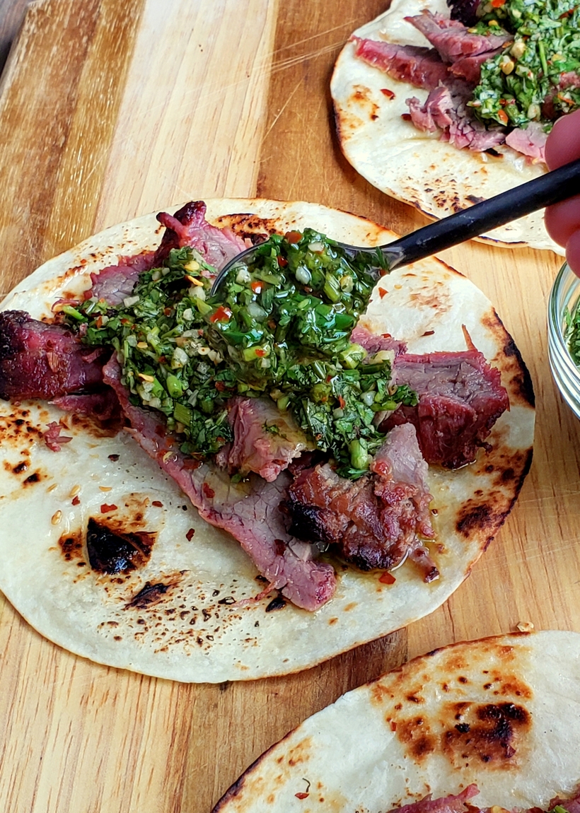 Make the best street food at home with these Flank Steak Tacos with Garlicky Cilantro Chimichurri. These tacos are essentially going to be your go-to summer, grilling meal. #noblepig #grilling #beef #flanksteak #chimichurri Chimichurri sauce being spooned on flank steak sitting on a tortilla.