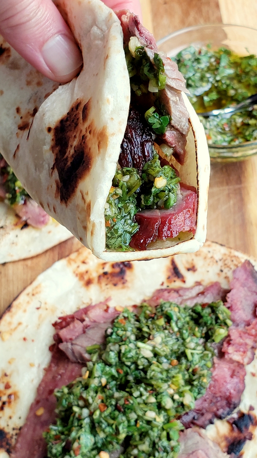 Make the best street food at home with these Flank Steak Tacos with Garlicky Cilantro Chimichurri. These tacos are essentially going to be your go-to summer, grilling meal. #noblepig #grilling #beef #flanksteak #chimichurri Hand holding a flank steak taco with one below..bowl of chimichurri in the background.