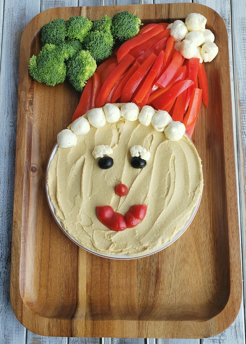 This Santa Snack Board is the perfect holiday centerpiece for every gathering this season. Not only is this healthy snack board festive and fun, it uses simple ingredients everyone loves. This Santa Board transports easily, feel free to take it along to every celebration this season. #noblepig #santasnackboard #diysantasnackboard #diy #healthysnackboard #christmas #christmasrecipes #easychristmasrecipes #holidayrecipes #santafood #santa #holidayentertaining #holidayfood #christmasappetizer Snack board being built to look like santa with hummus face, broccoli, red peppers and cheese.