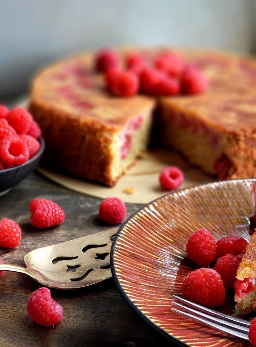 Cake with a slice missing surrounded by raspberries.