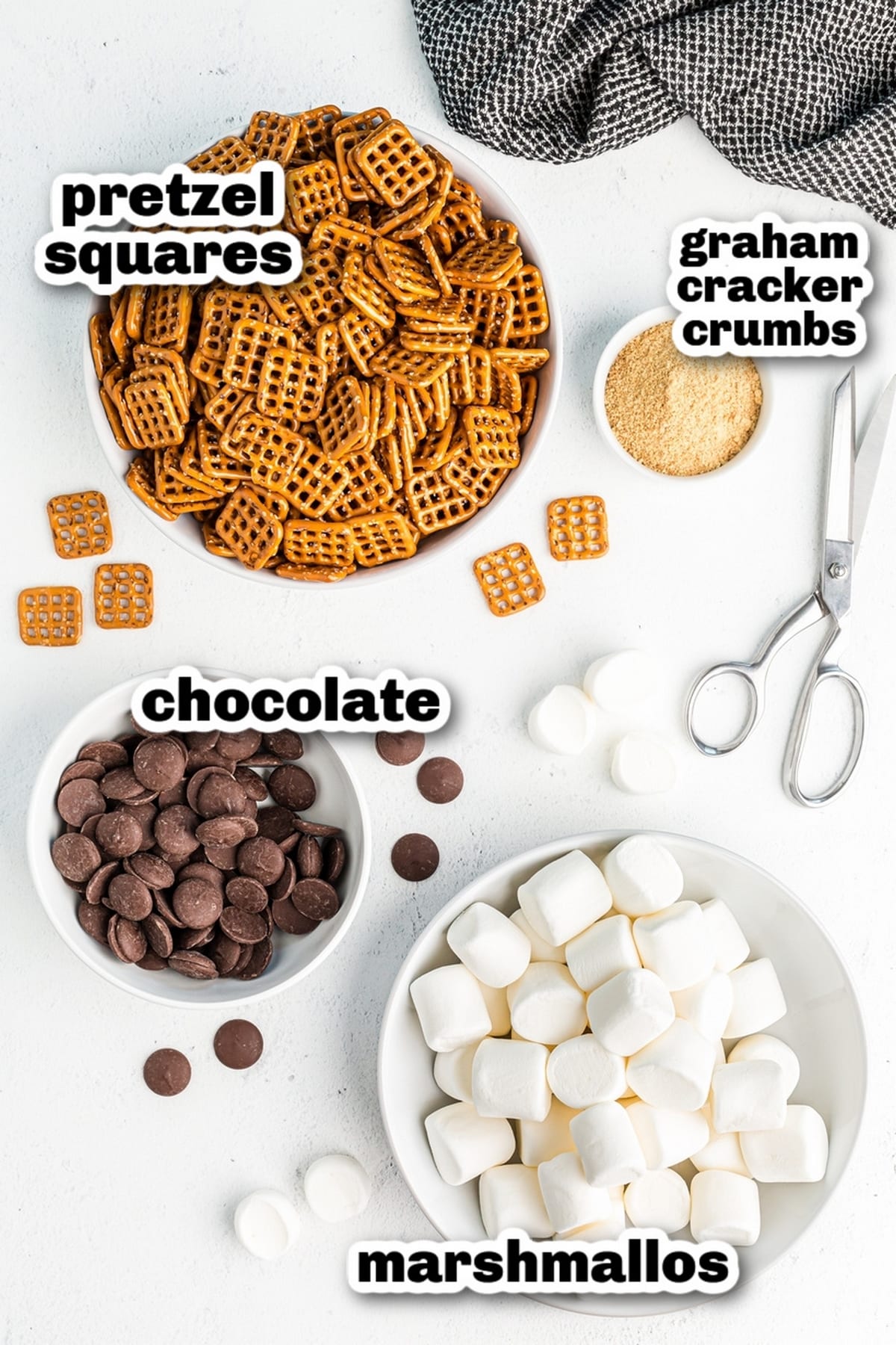 s’mores pretzels ingredients with chocolate, marshmallows, pretzels, and graham cracker crumbs Ingredients for s’mores pretzels including square pretzels, chocolate melting wafers, graham cracker crumbs, and marshmallows arranged in bowls