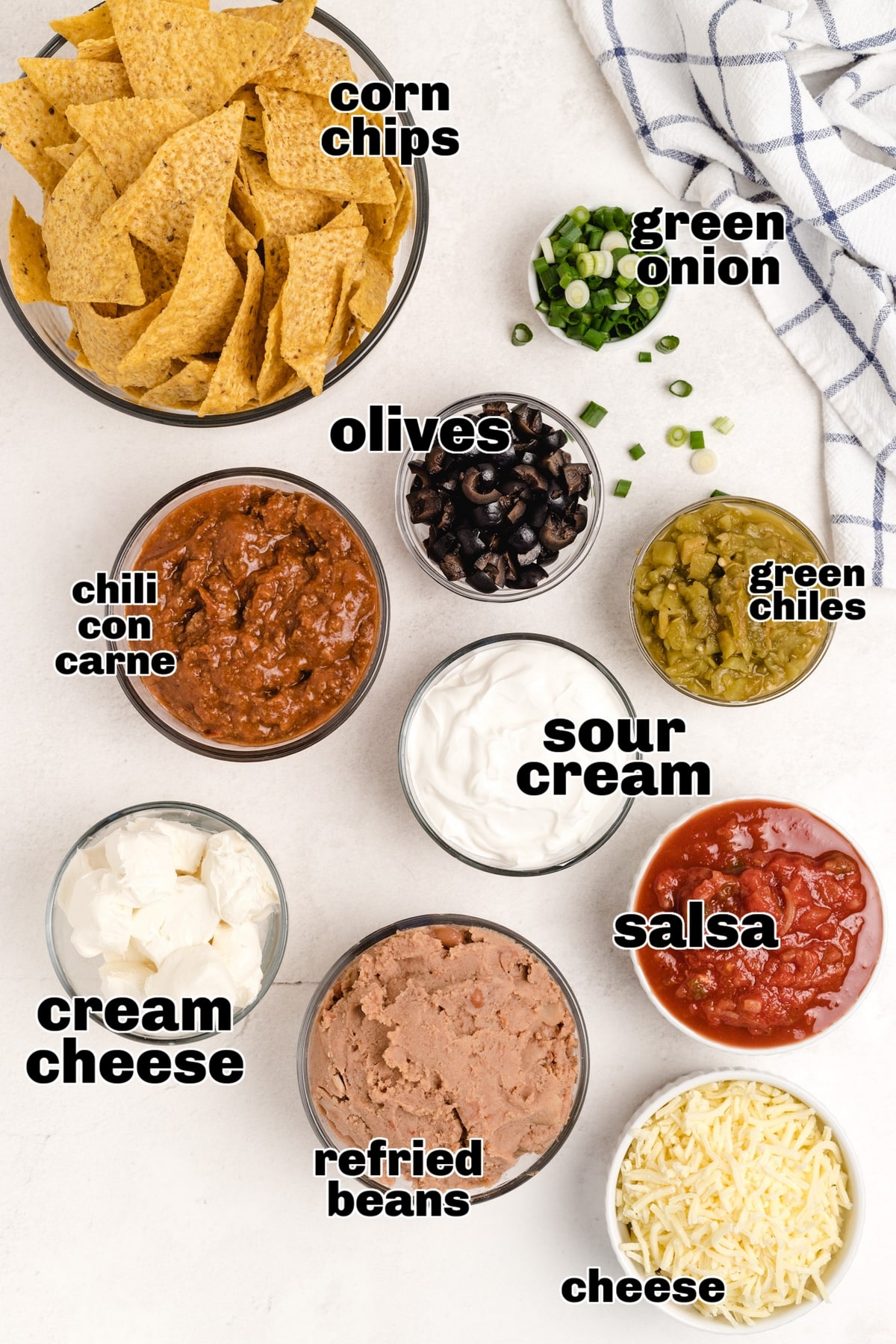 Ingredients for baked taco dip arranged on a countertop: refried beans, chili con carne, salsa, olives, cheese, and green onions.