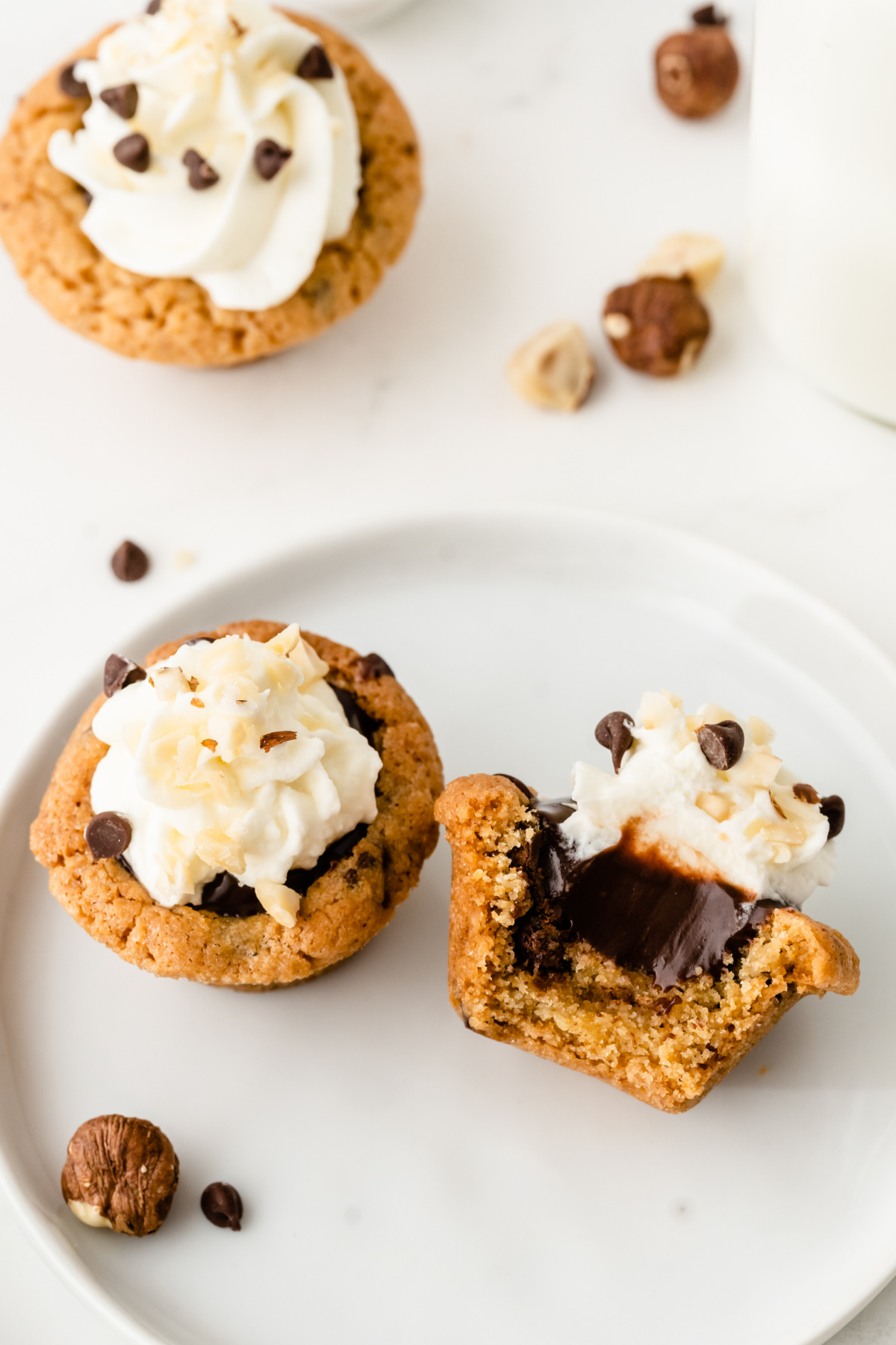 Cookie cup dessert filled with Nutella ganache and topped with whipped cream, shown whole and split open Chocolate chip cookie cup with Nutella center and whipped cream topping, one broken open to show filling