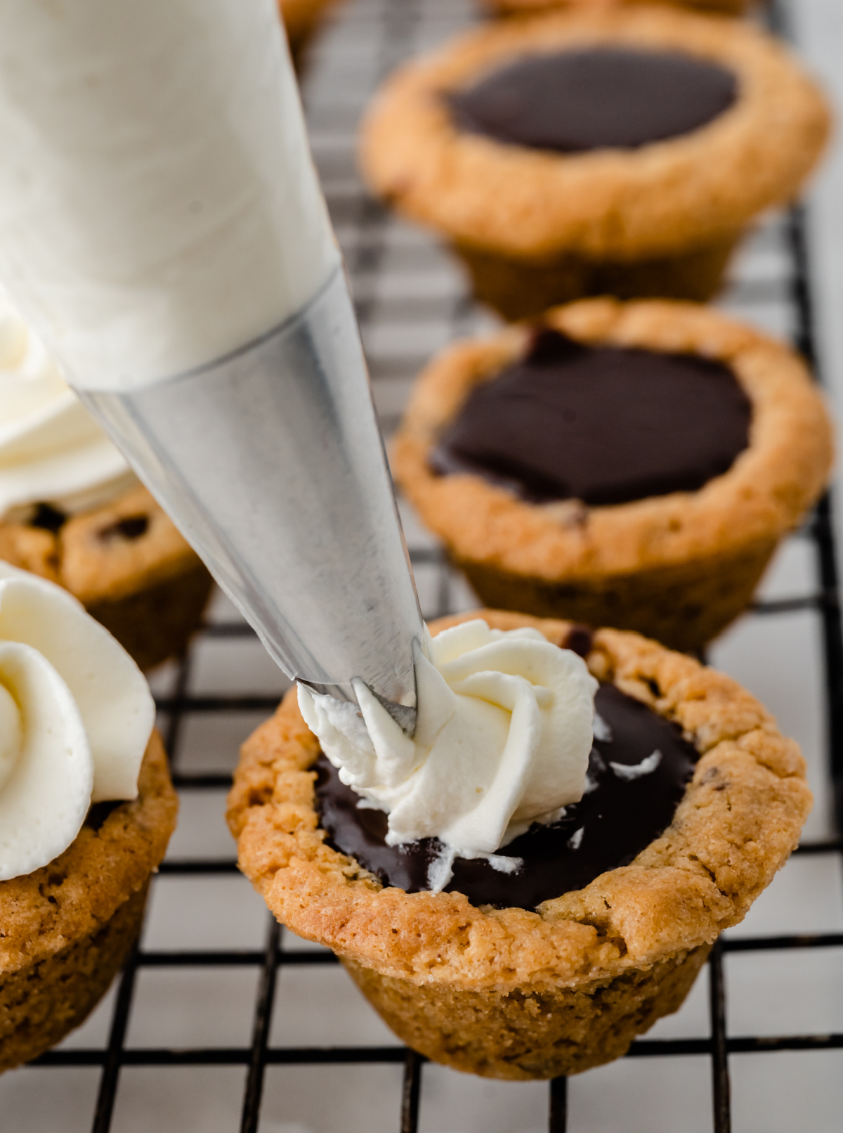 Nutella chocolate cookie cups with a set Nutella center finished with piped whipped cream Nutella chocolate cookie cups being topped with whipped cream on a cooling rack