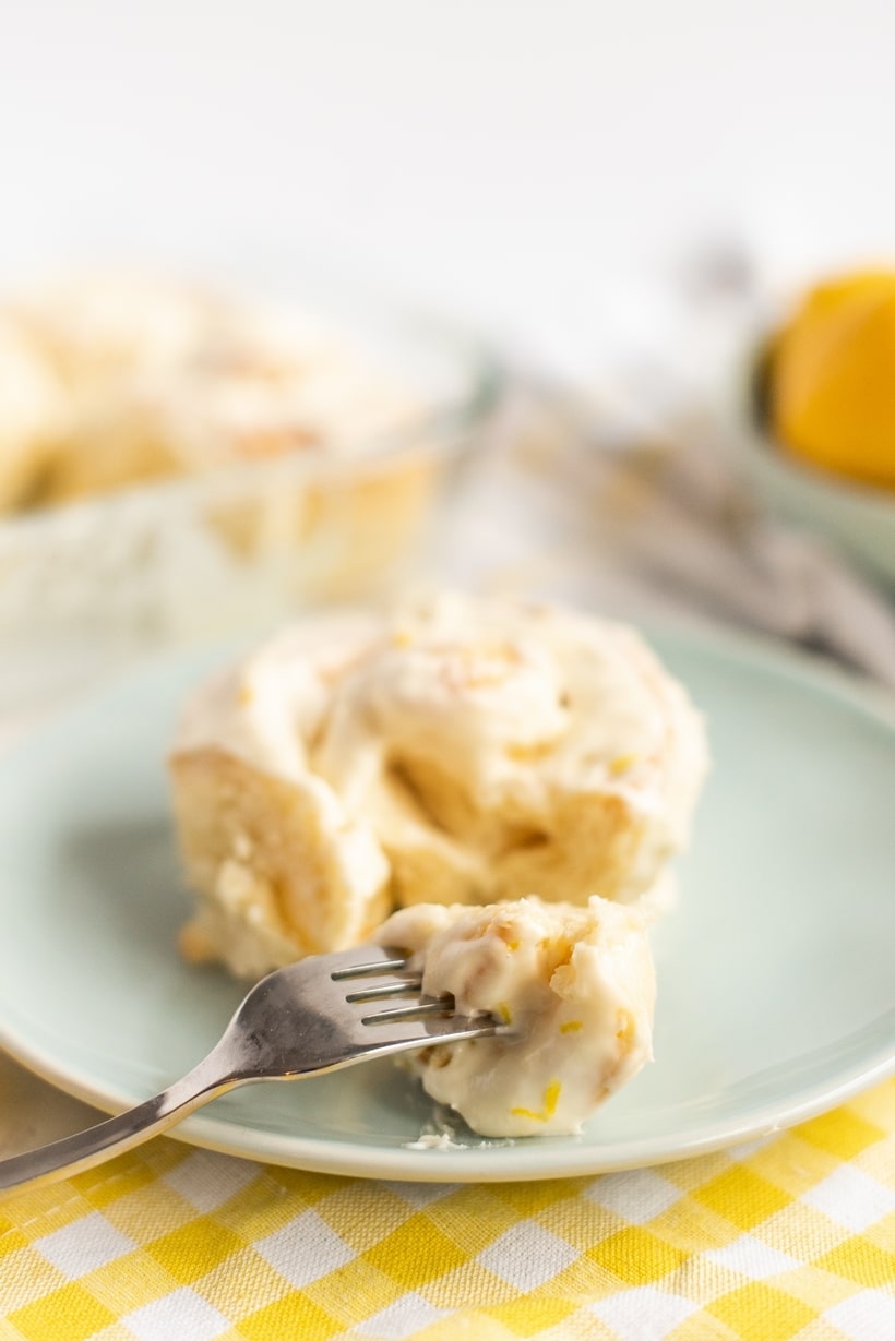 A soft, fluffy lemon bun with cream cheese icing on a white plate, with a fork lifting a bite-sized piece.