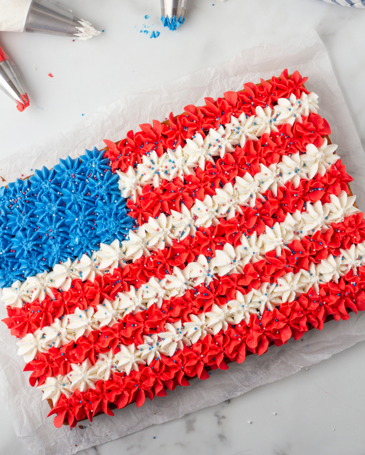  Overhead view of buttercream American flag cake decorated with piped red, white, and blue stars, piping bags visible in the background.