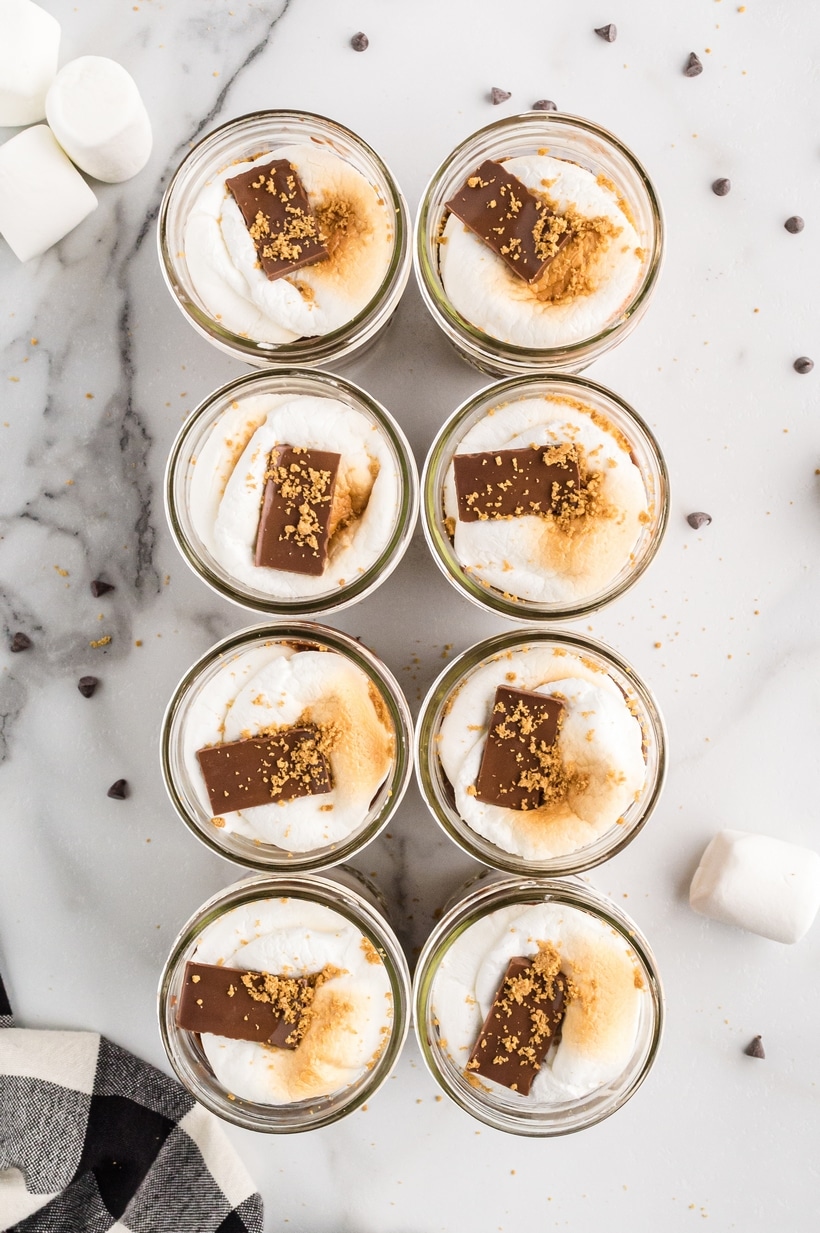 Overhead view of multiple s’mores parfaits with chocolate and graham topping Top-down view of eight s’mores pudding parfaits in jars, each topped with toasted marshmallow and chocolate