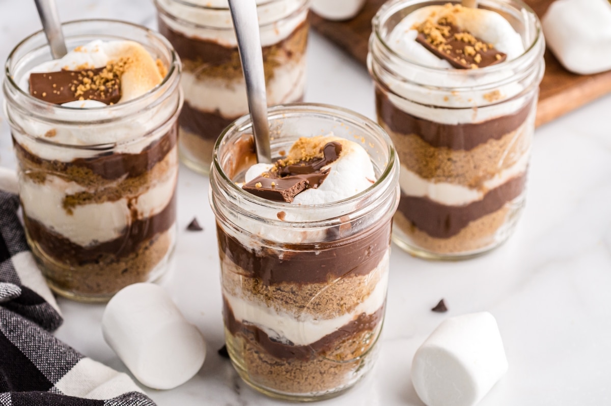 S’mores dessert cups with layers of chocolate pudding and whipped marshmallow Multiple no-bake s’mores dessert jars on a white surface with chocolate, graham cracker, and marshmallow layers
