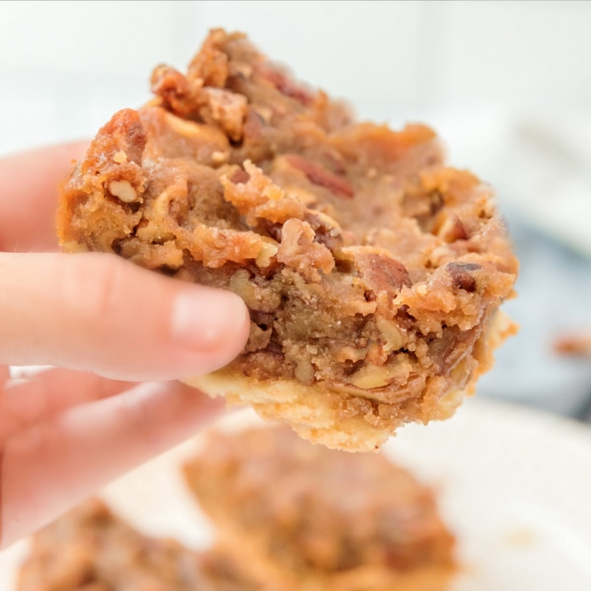 Pecan Pie Bar Held in Hand Hand holding a pecan pie bar, showcasing its rich pecan filling and buttery shortbread crust.
