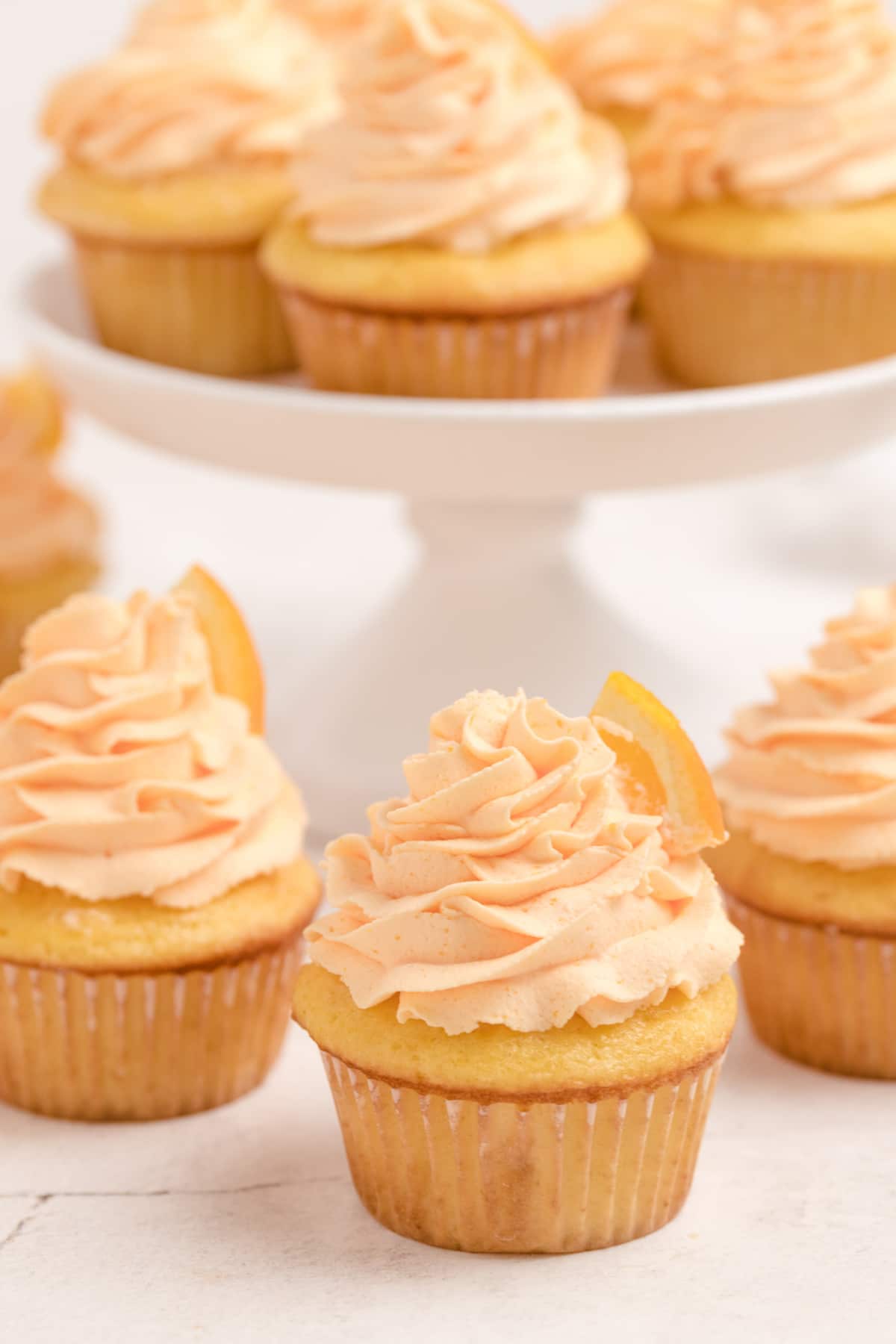 Orange Cupcakes with Whipped Orange Frosting with orange slices A close-up of a single cupcake with piped frosting, with a cake stand full of cupcakes slightly blurred in the background.