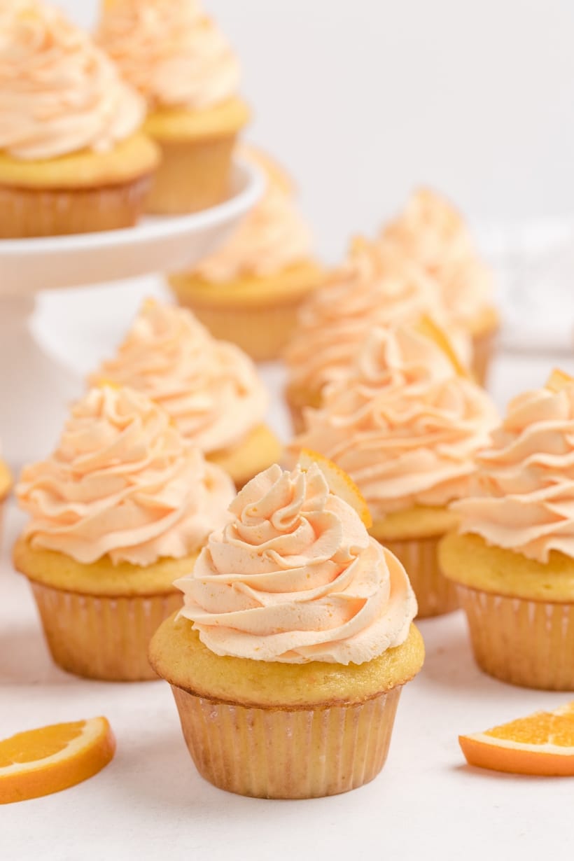 Dreamy Orange Cupcakes with Whipped Orange Frosting on a cake stand A close-up of freshly frosted Dreamy Orange Cupcakes with Whipped Orange Frosting, with more cupcakes displayed on a cake stand in the background.
