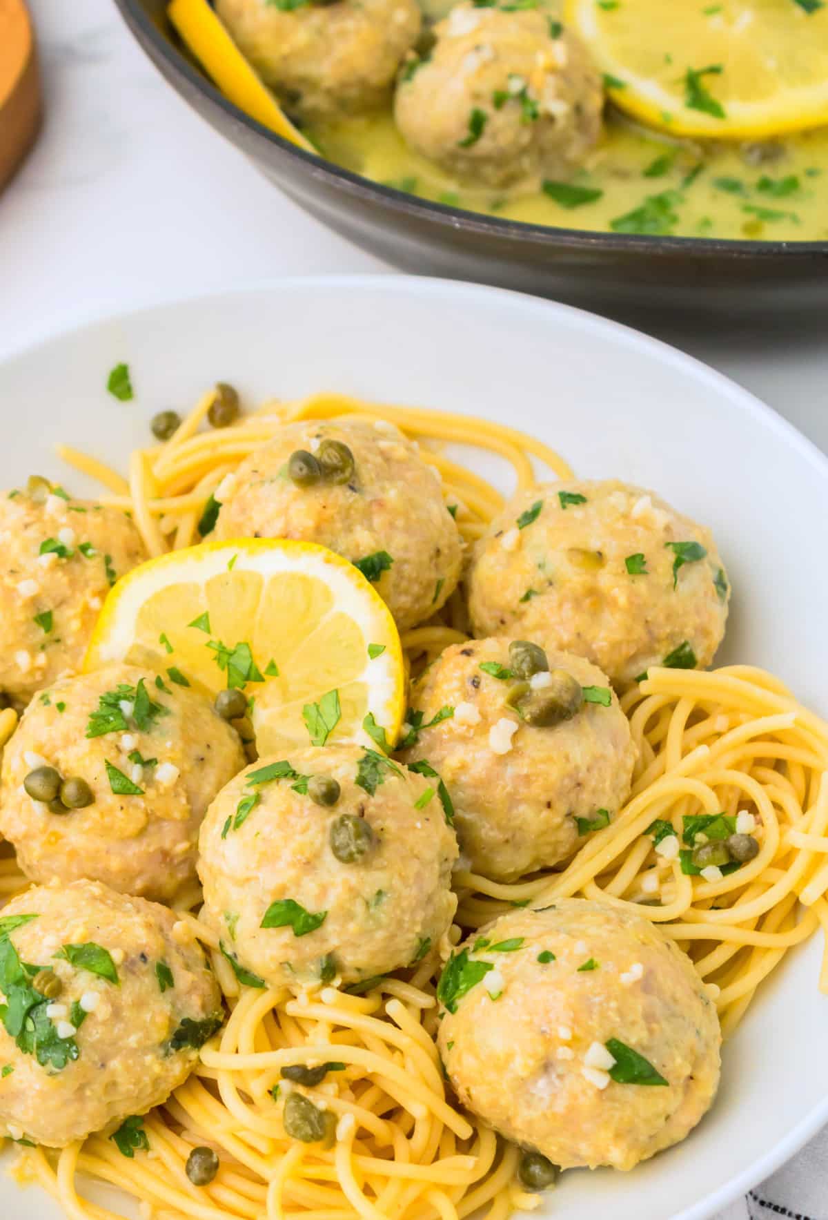 plated chicken piccata meatballs with skillet in background Plate of chicken piccata meatballs over pasta with skillet of extra meatballs in the background