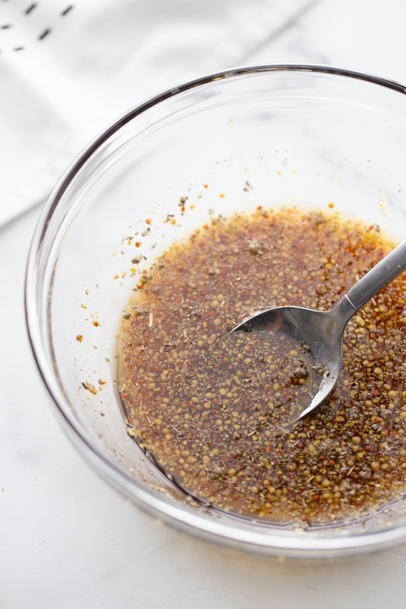 Mixing maple syrup, mustard, and sage for a flavorful sausage glaze. A bowl of maple syrup, mustard, and crushed sage being stirred together to make a savory glaze.