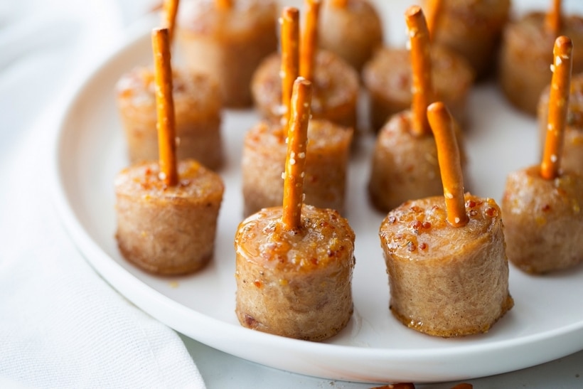Finished maple-glazed sausage bites served on a white platter with pretzel skewers. sMaple-glazed sausage bites skewered with pretzel sticks arranged on a white platter.