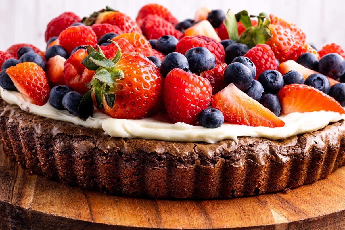 chocolate brownie fruit pizza with berries and thick cream cheese layer Side view of chocolate brownie pizza topped with cream cheese frosting and fruit