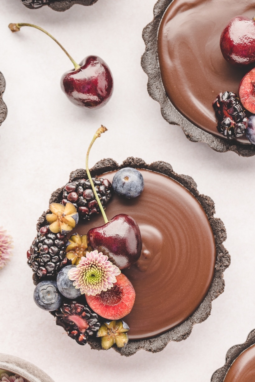 Mini Chocolate Ganache Tarts Top-down view of a mini chocolate ganache tart topped with fresh berries and edible flowers on a white background.
