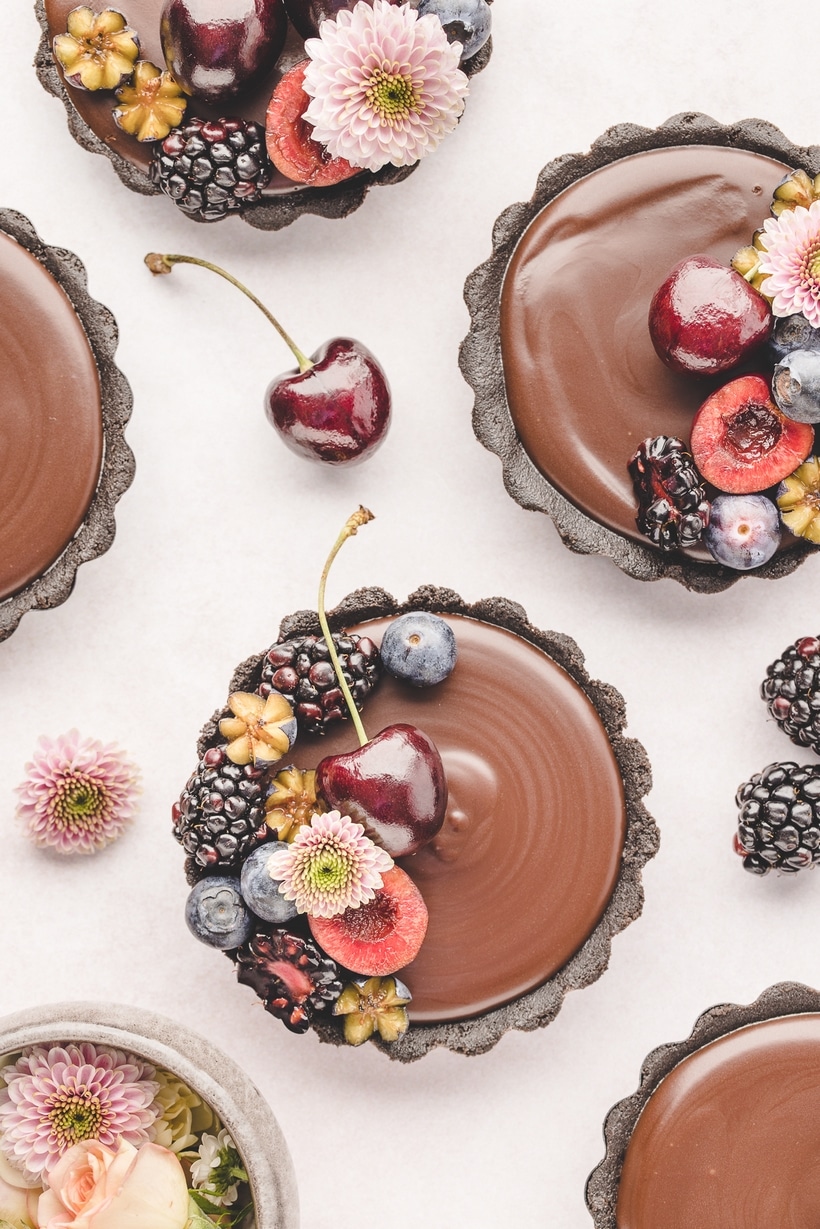 embellished with fruit Mini Chocolate Ganache Tarts Top-down photo of multiple mini chocolate ganache tarts arranged on a surface, some decorated and some plain.