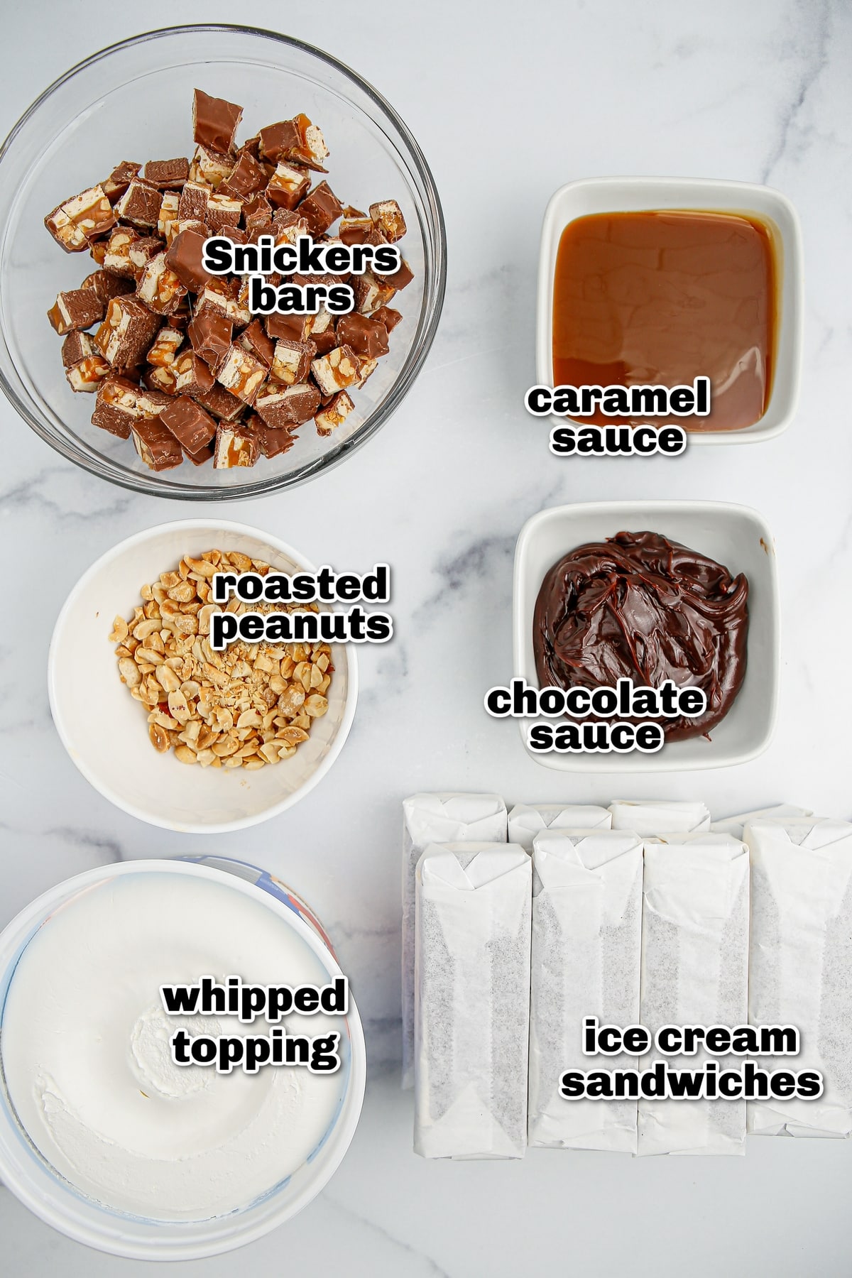 Overhead view of ingredients for snickers ice cream cake including whipped topping, chocolate and caramel sauces, peanuts, Snickers bars, and ice cream sandwiches