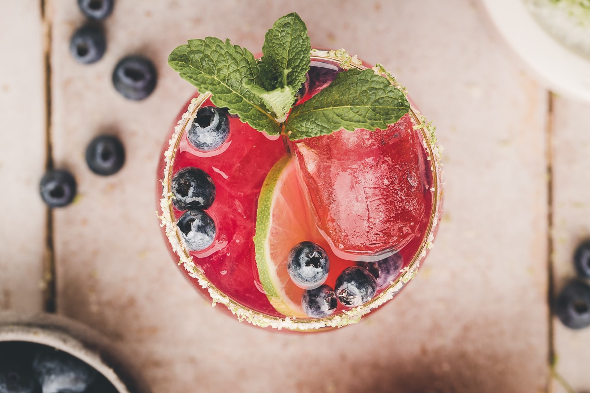 Blueberry Margarita top view Overhead view of a blueberry margarita filled with ice, lime slices, and blueberries.