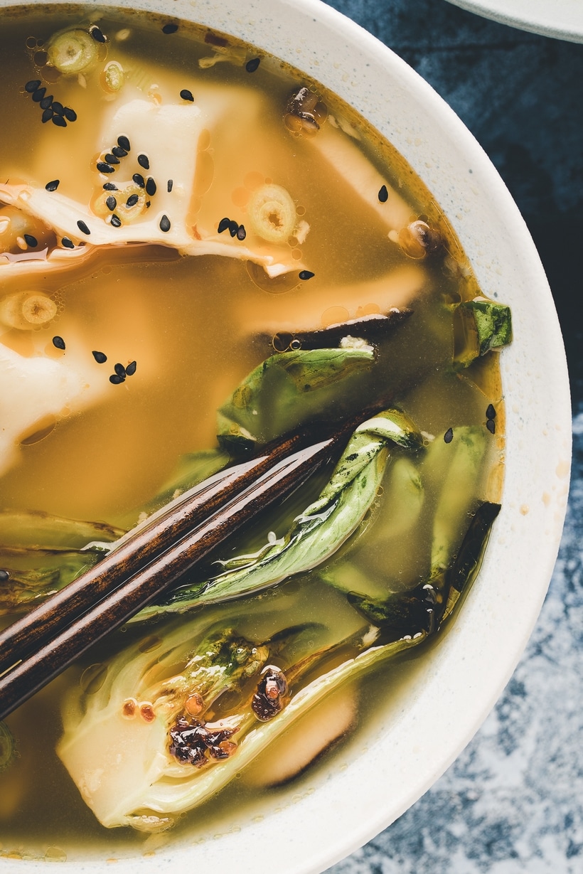 Close-Up of Wonton Soup Bowl with Chopsticks Close-up of a steaming bowl of wonton soup with tender wontons, bok choy, and garnishes of scallions, sesame seeds, and chili oil, served with chopsticks resting on the bowl.