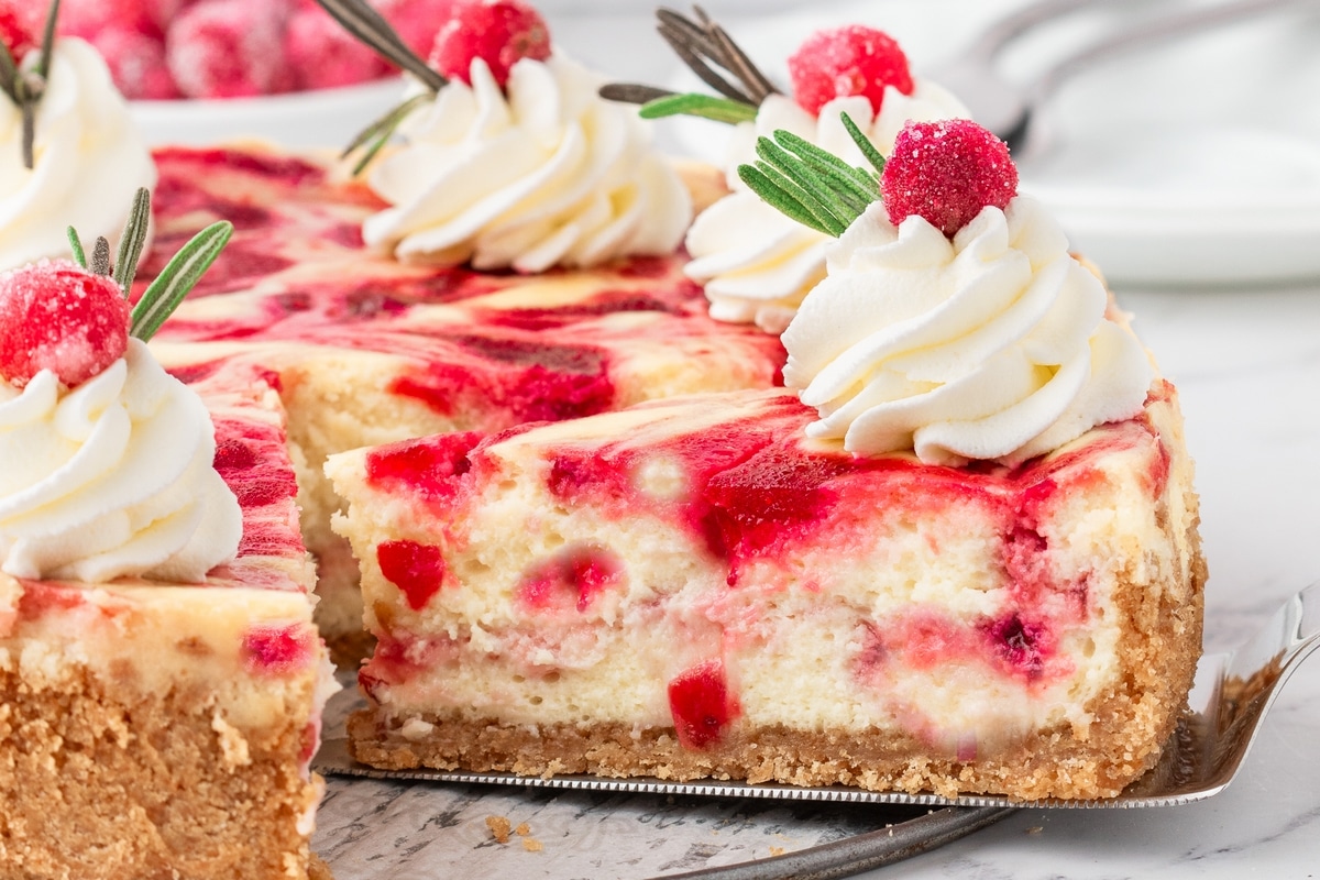 A slice of cranberry cheesecake being lifted from the whole cheesecake, revealing the cranberry swirl and creamy texture.