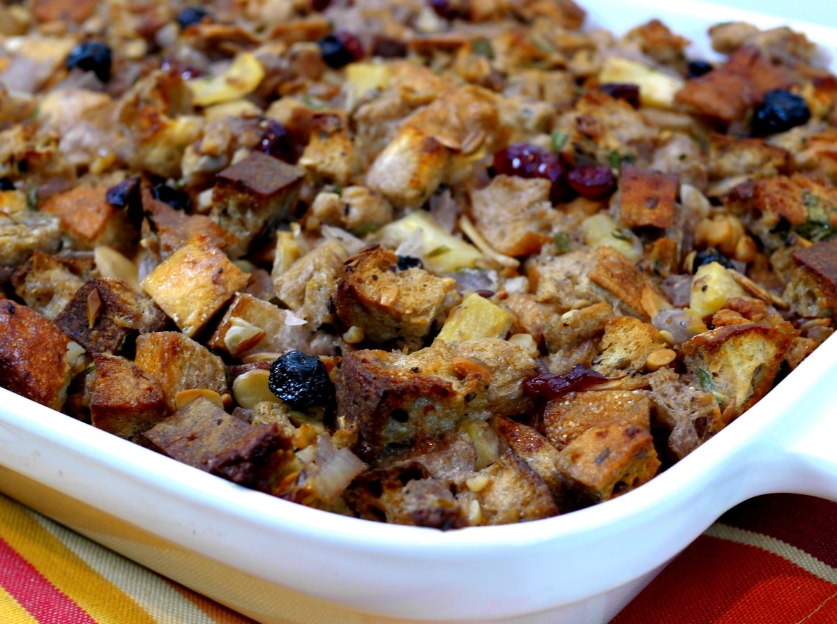 Top view of baked rustic multigrain stuffing with nuts and dried fruits in white dish Overhead view of rustic multigrain stuffing with nuts and dried fruits in a white baking dish, showcasing its golden-brown crust and hearty texture.