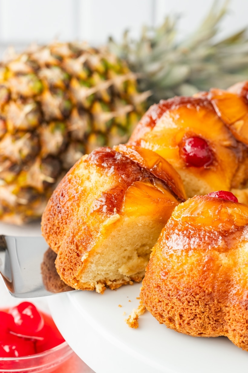 Pineapple upside down Bundt cake being lifted from a cake stand, with a whole pineapple in the background.