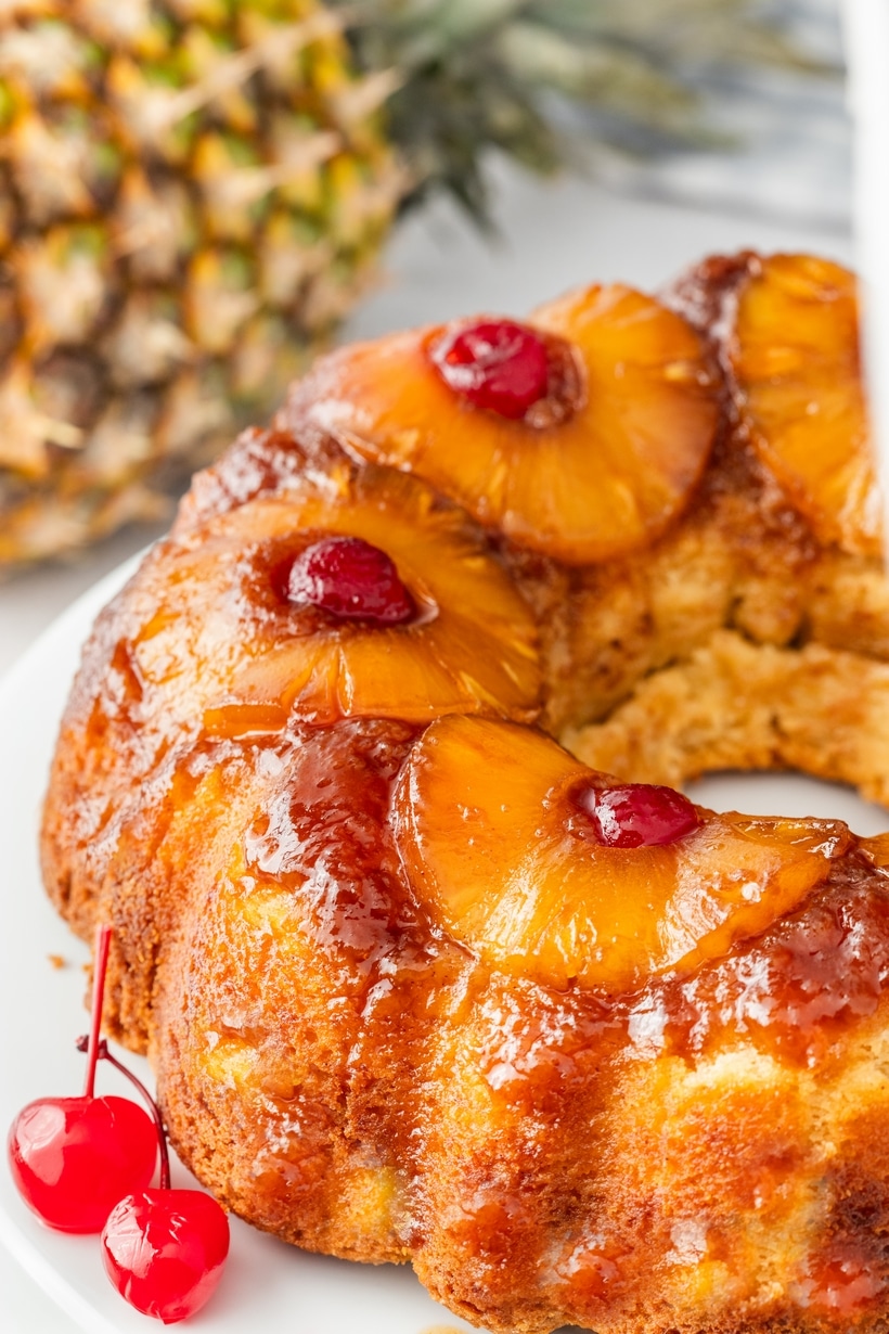 Side view of pineapple upside down cake with glossy pineapple and cherry topping, whole pineapple in background.