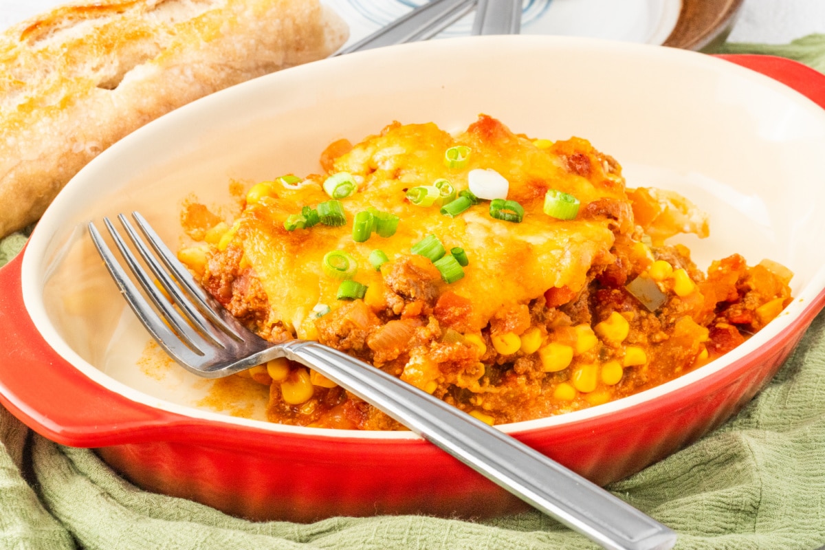 Ground beef casserole with pasta, corn, and cheese in a red baking dish Cheesy ground beef casserole served with bread and topped with green onions