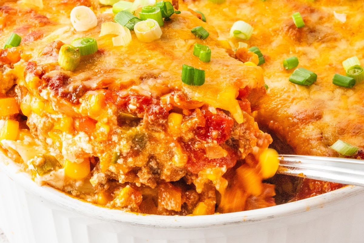 Scooping a serving of cheesy ground beef pasta bake from casserole dish Serving being scooped from baked ground beef casserole with corn, pasta, and cheddar
