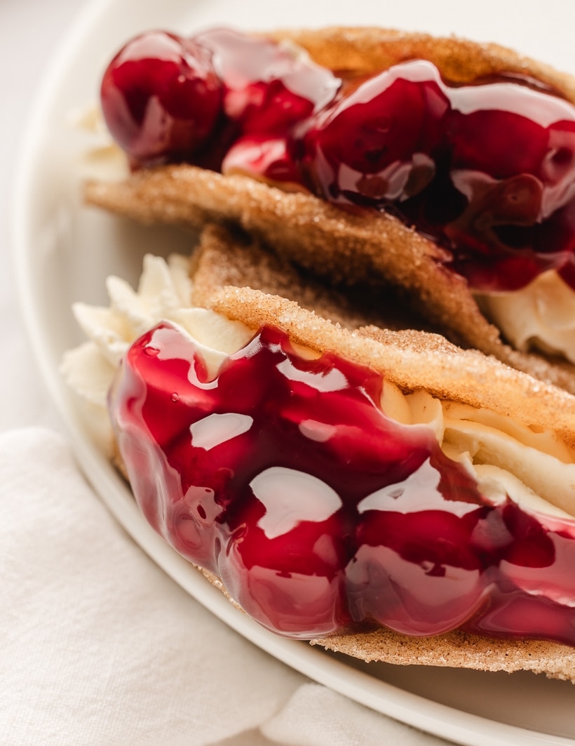 Cherry Cheesecake Tacos on White Plate Top view of two cherry cheesecake tacos on a white plate, filled with cheesecake and topped with cherry pie filling.